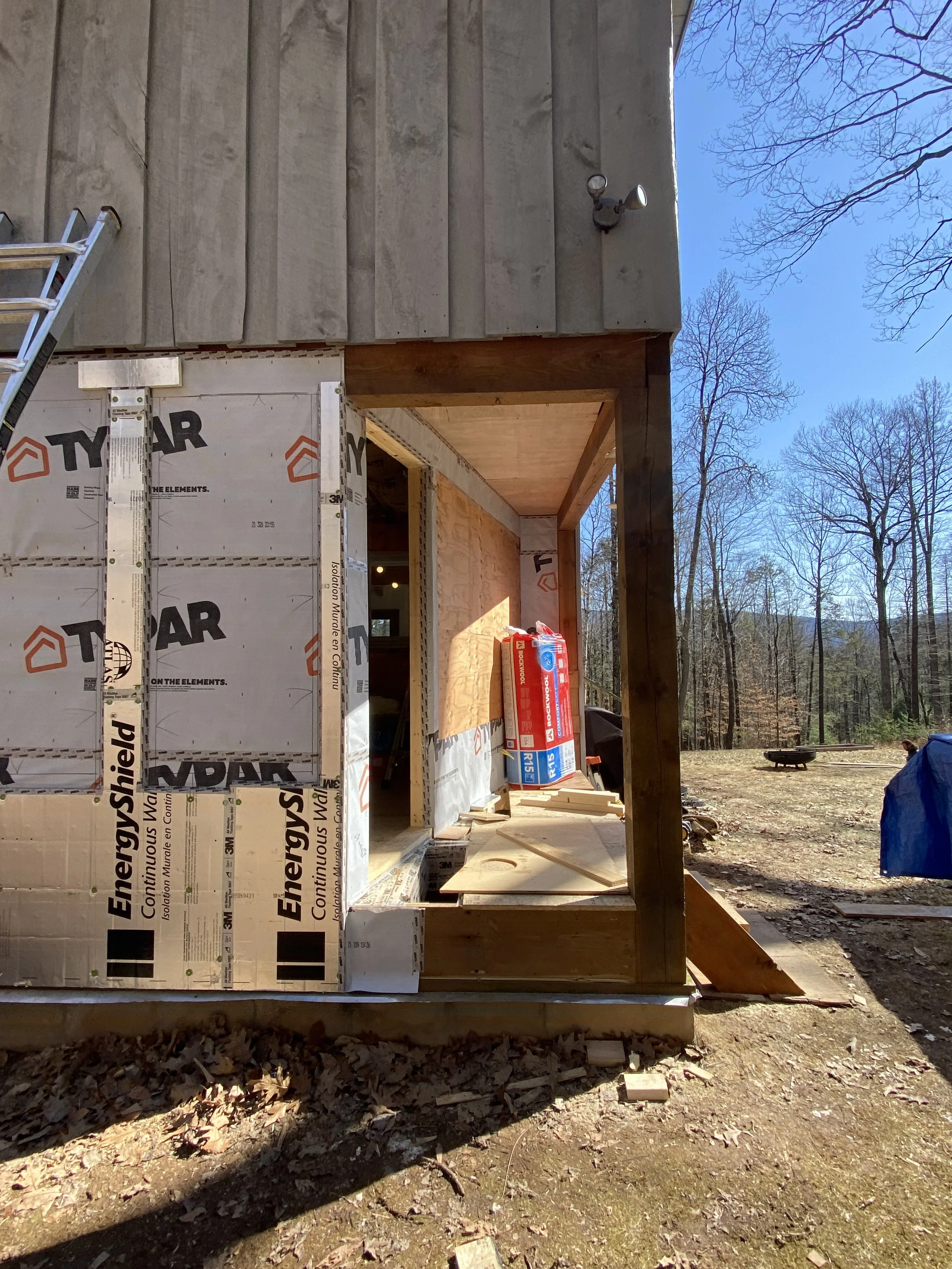 Construction site of a house with wooden framing and insulation, surrounded by trees, with construction materials and tools visible.