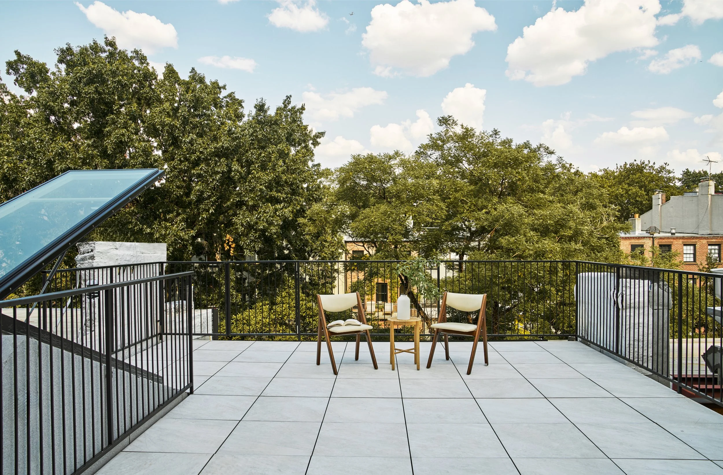 A rooftop terrace with two chairs, a small table with a vase and plant, and a view of trees and buildings under a partly cloudy sky.