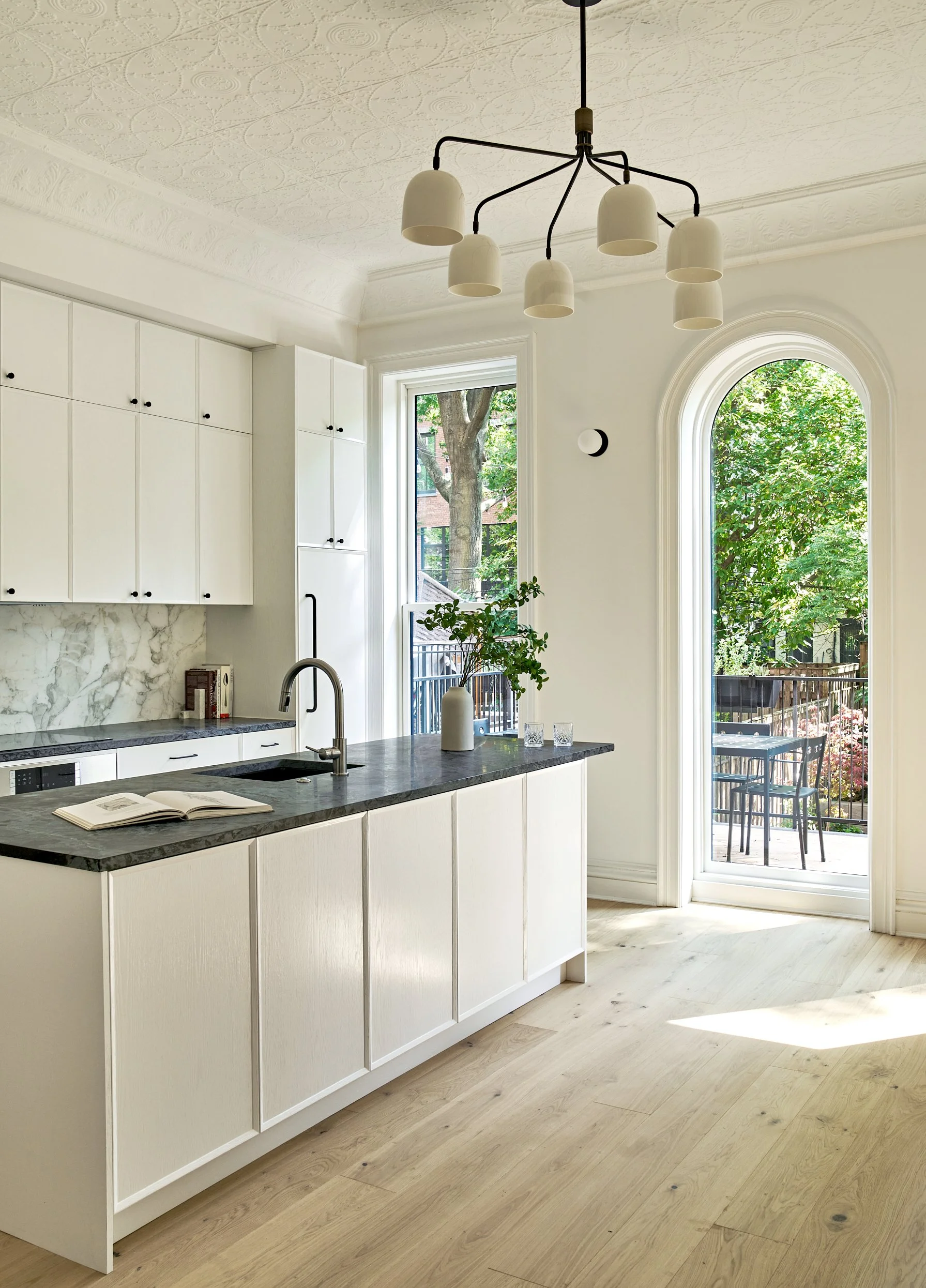 Bright, modern kitchen with white cabinets, a black and white marble backsplash, and a black countertop island. There is an open book on the island, a faucet, and a potted plant on the countertop. Large windows and a glass door offer views of a sunny outdoor patio with outdoor furniture and greenery.