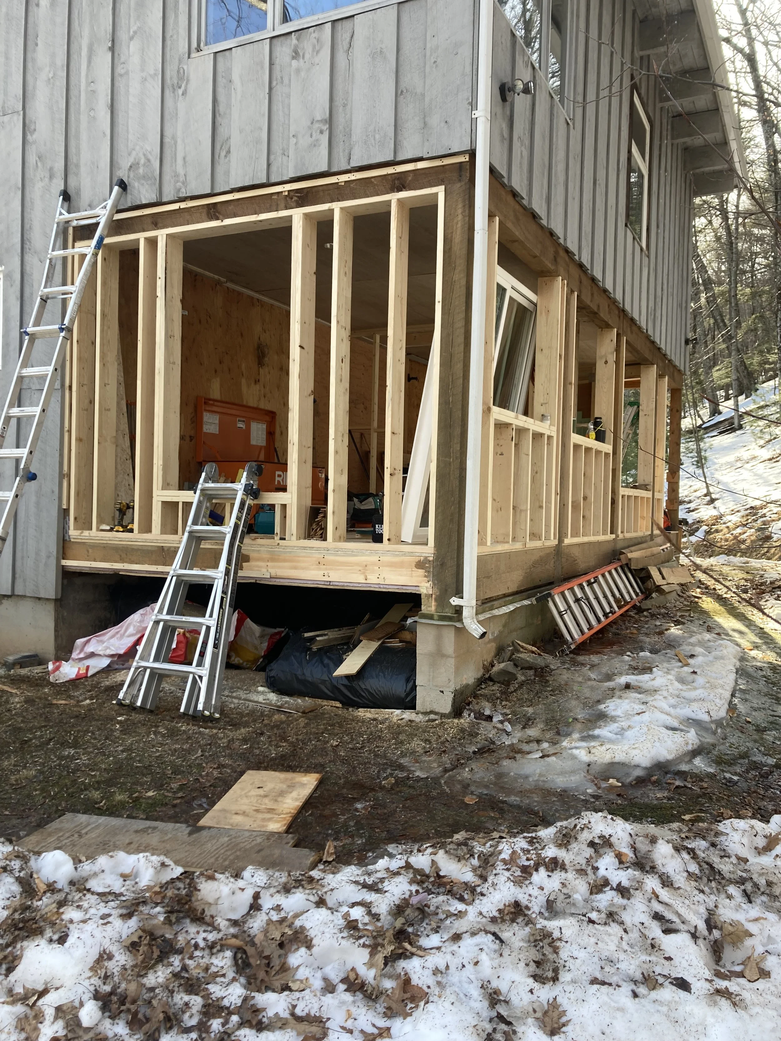 Construction site of a house with a wooden frame and unfinished exterior, surrounded by snow and leaves.
