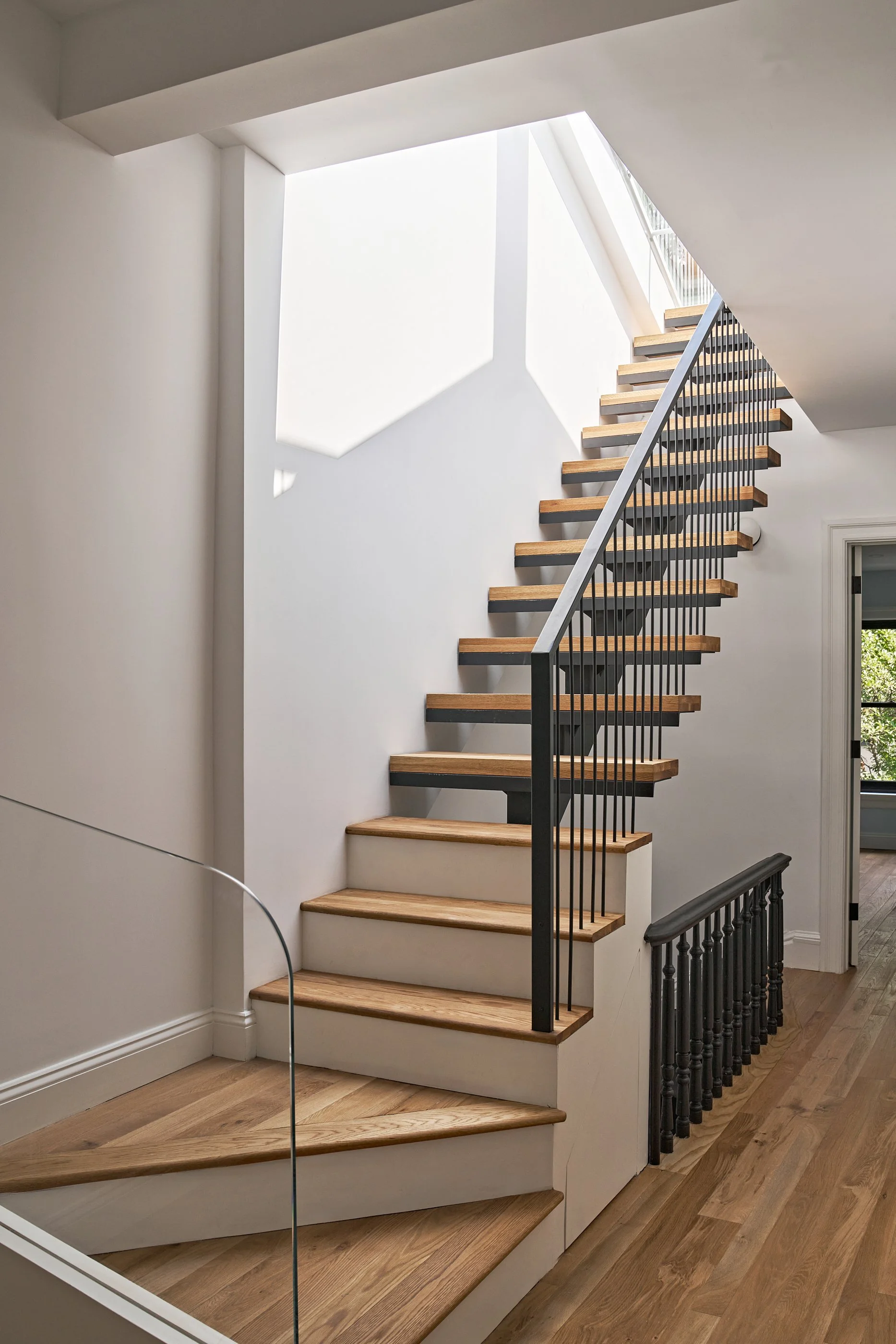 Interior view of a staircase with wooden steps, black metal railing, and a large window letting in natural light, with hardwood flooring.
