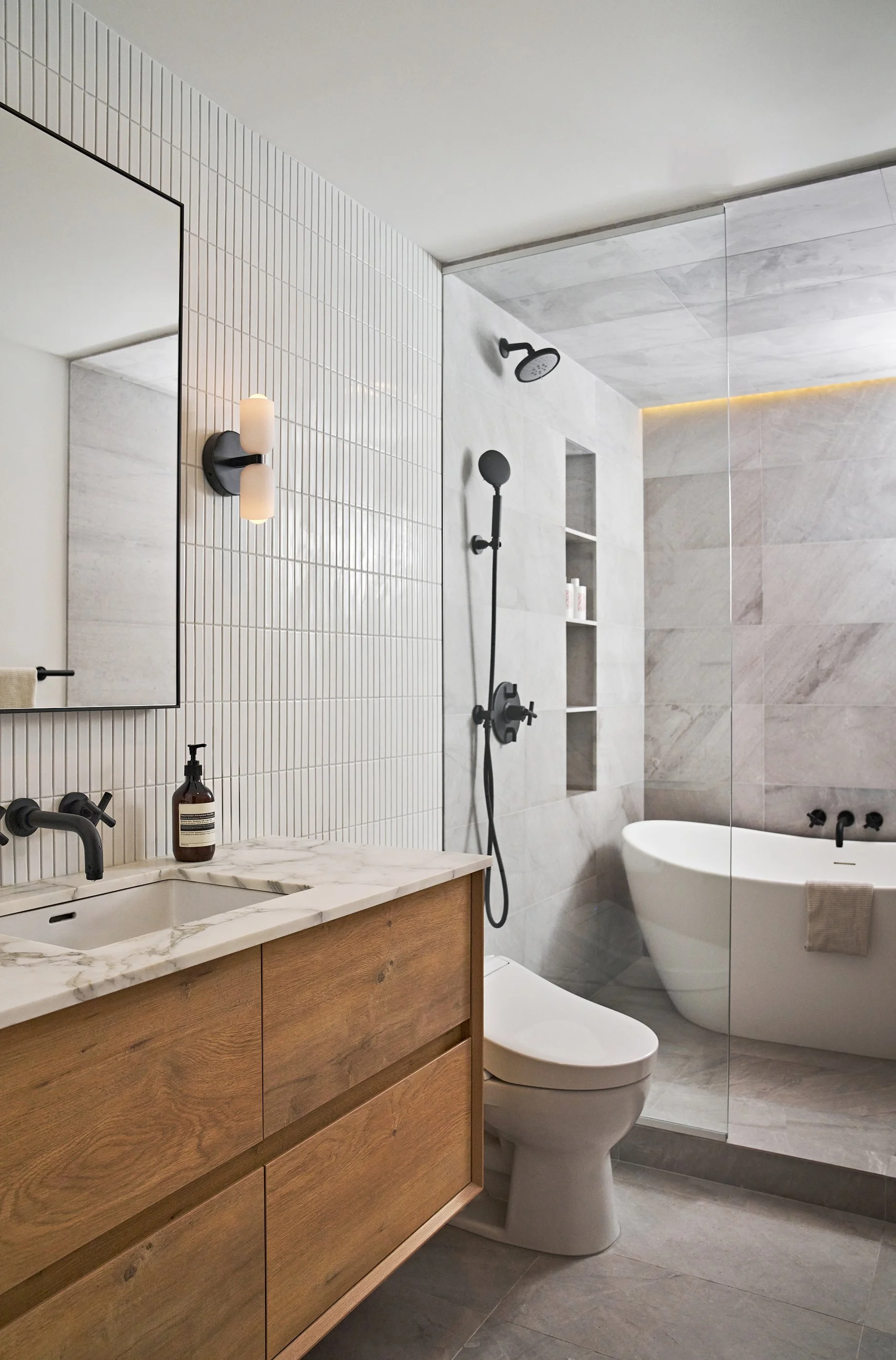 Modern bathroom with white tiled walls, wooden vanity with marble countertop, black fixtures, a toilet, and a standalone bathtub.