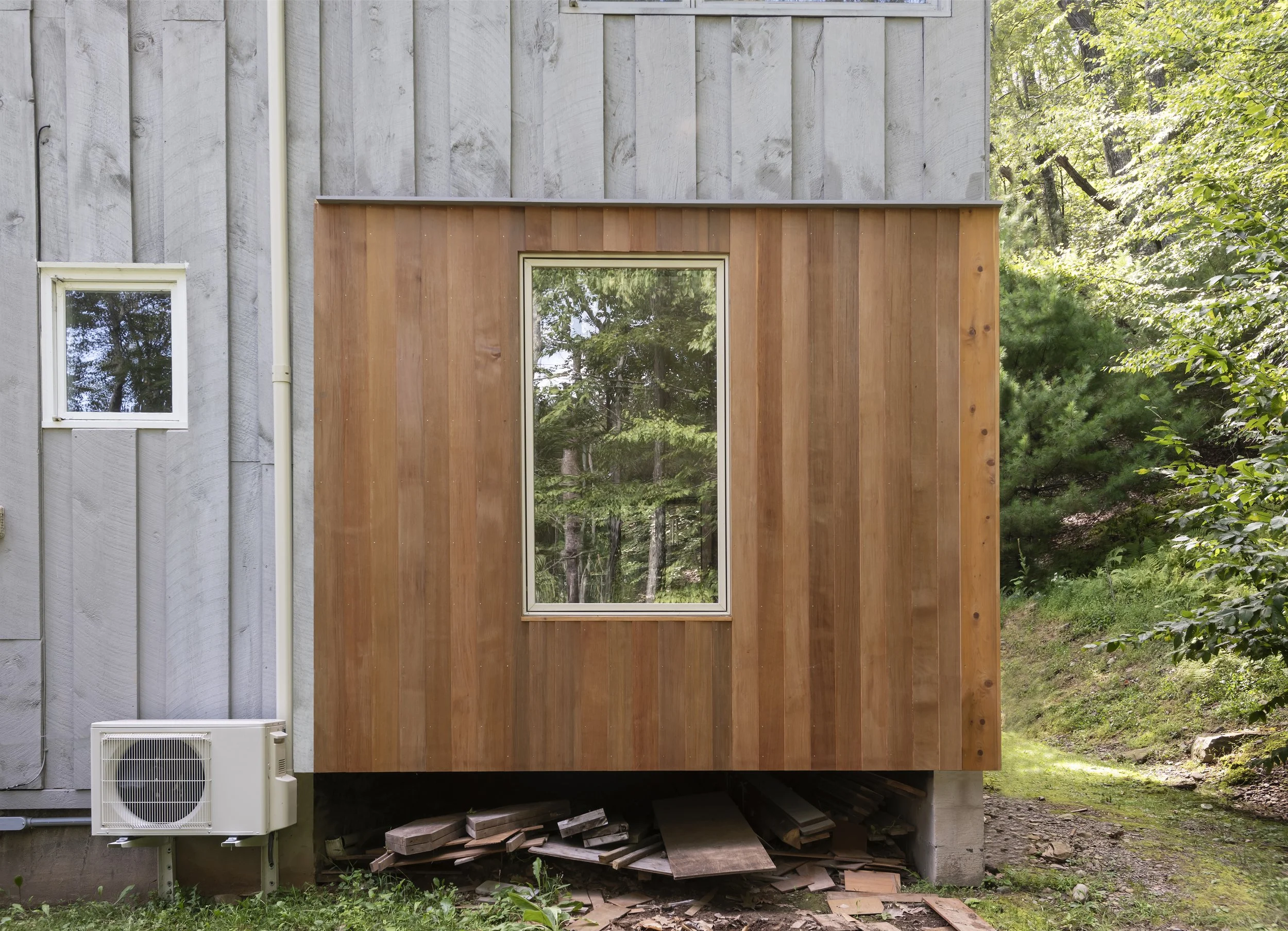 The back exterior of a house showing a small, new wooden addition with vertical siding and a large center window. The house has gray vertical siding, a small window on the left, and an outdoor AC unit below the smaller window. Piled wood and debris are underneath the addition, and there is a forested area in the background.
