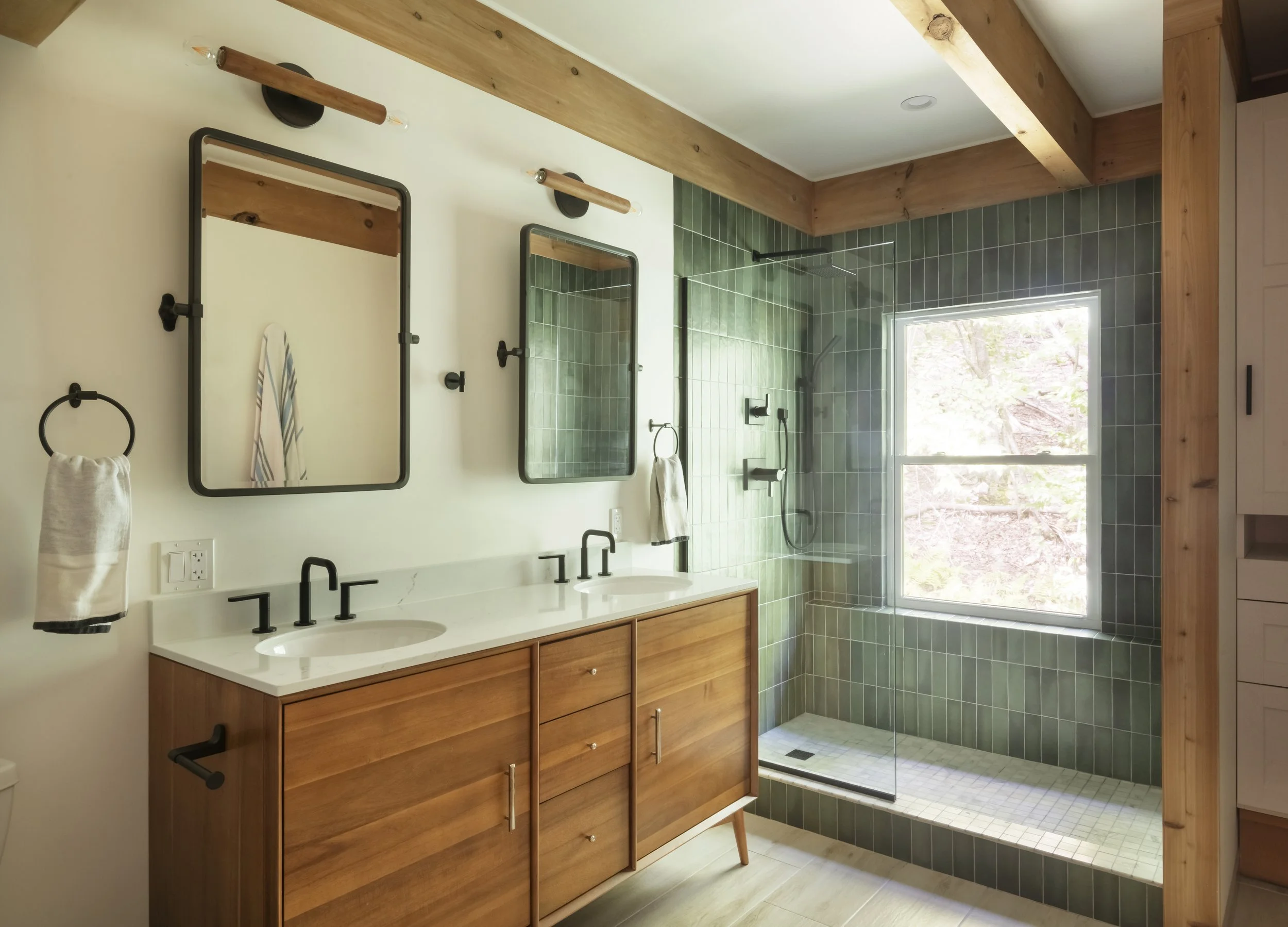 Modern bathroom featuring a wooden vanity with two sinks, black fixtures, two mirrors, wall-mounted towels, green tiled shower area with a window, and wood accents on the ceiling and shower enclosure