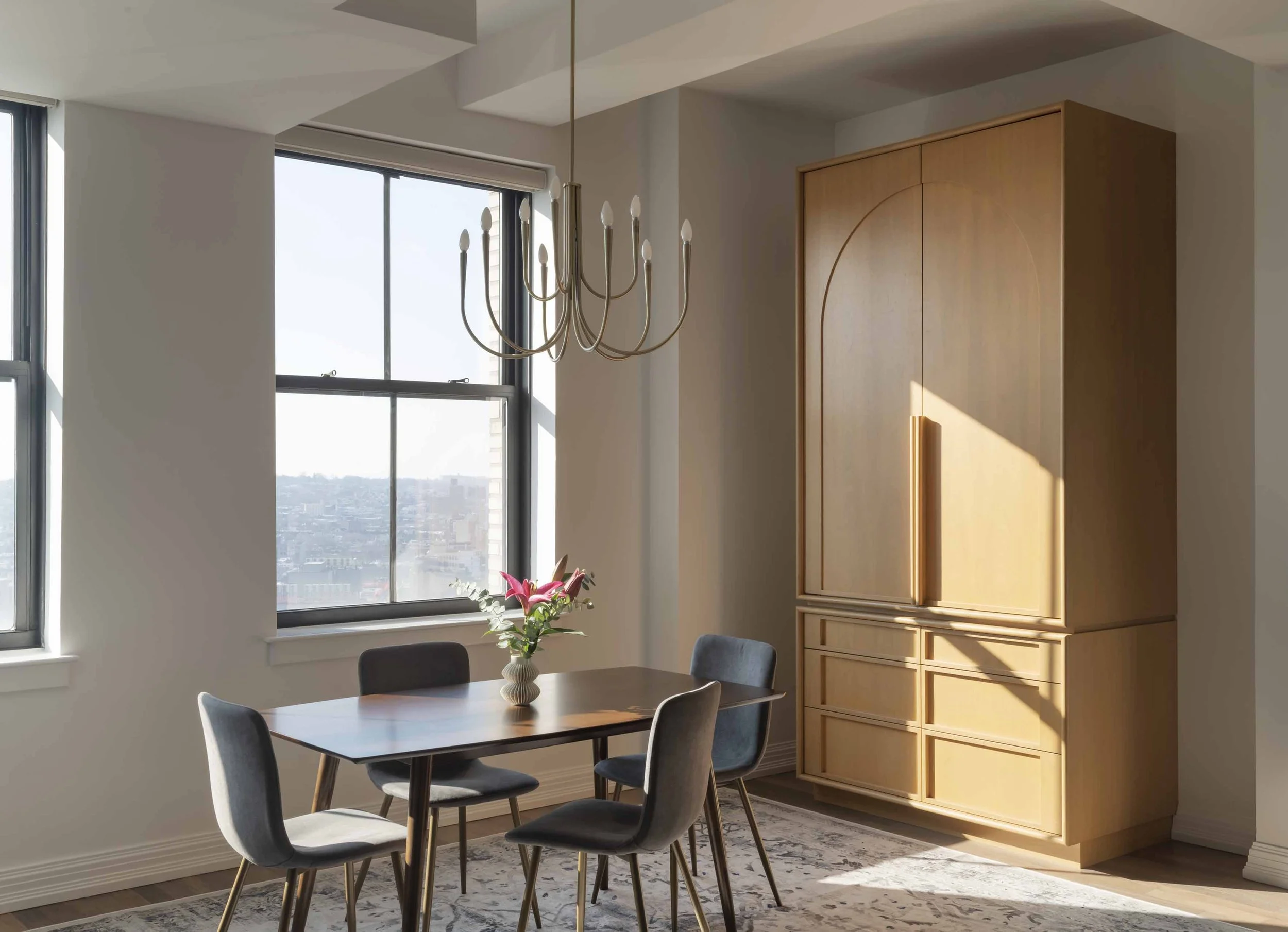 A dining area with a wooden table, gray upholstered chairs, a vase with pink flowers, a window with a city view, a modern chandelier, and a large wooden cabinet in a bright room.