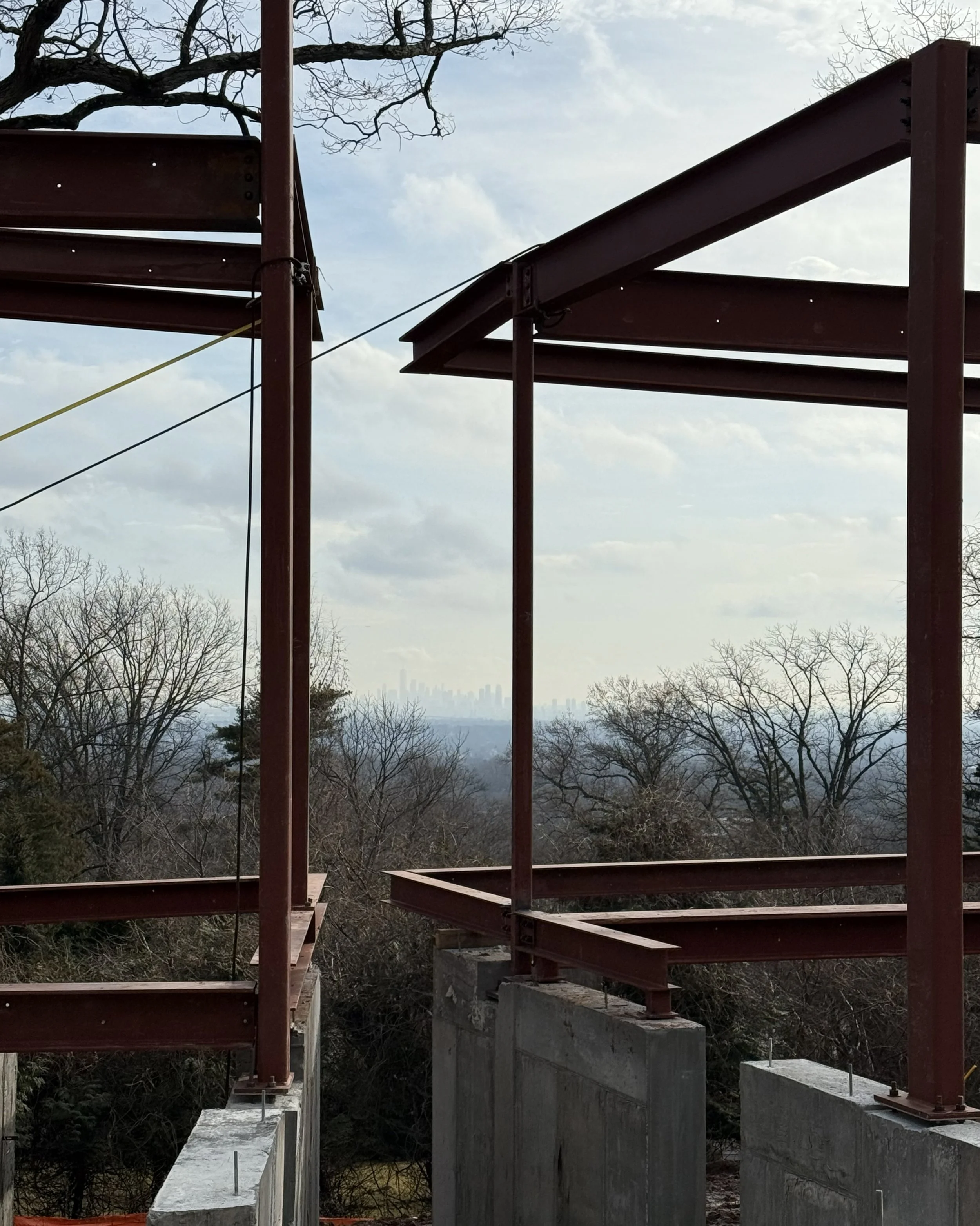 Construction site with red steel framework and concrete supports for a bridge, with trees and cloudy sky in the background.