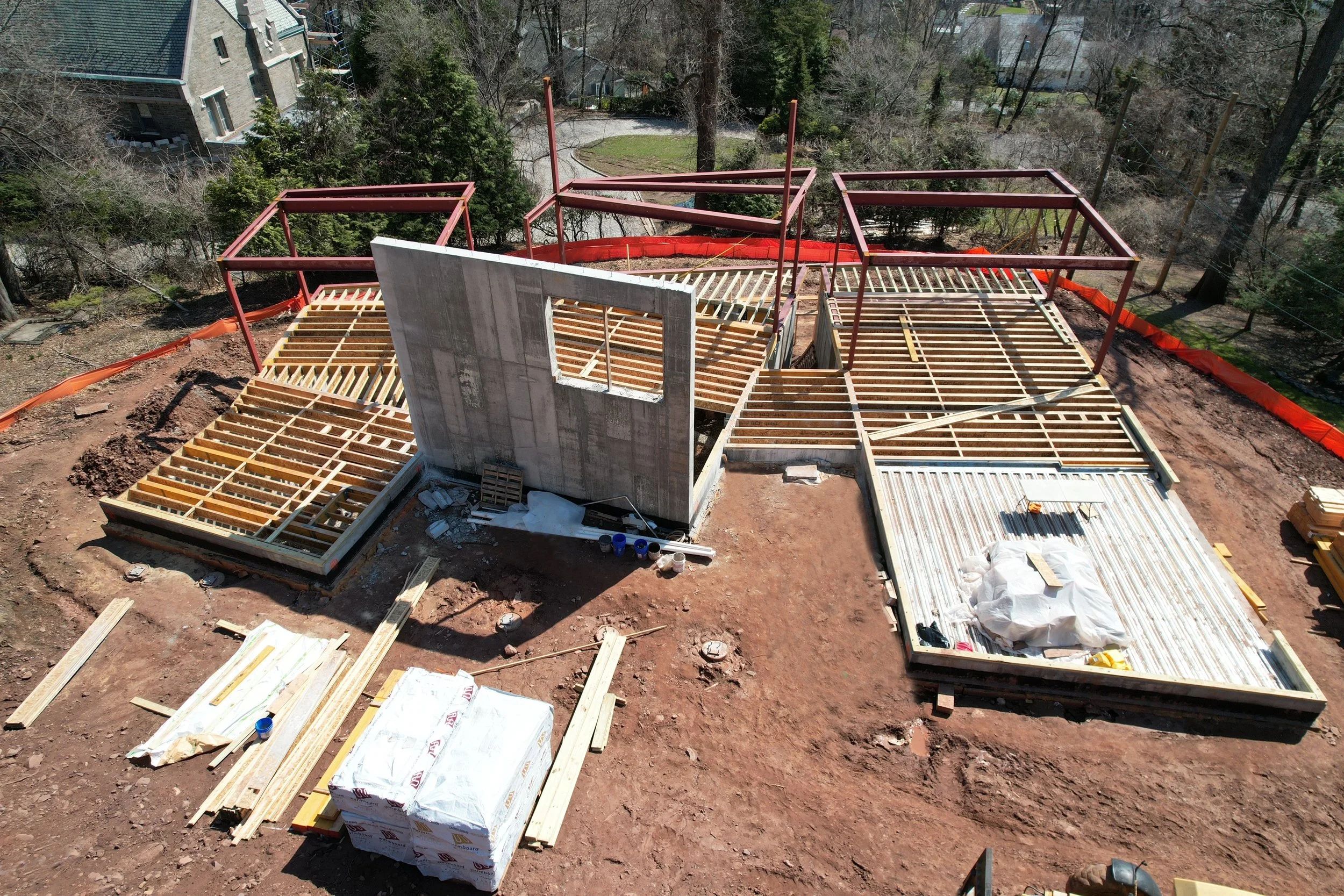 Construction site with wooden framing for a building, including a concrete wall with a window opening, surrounded by trees and residential houses.
