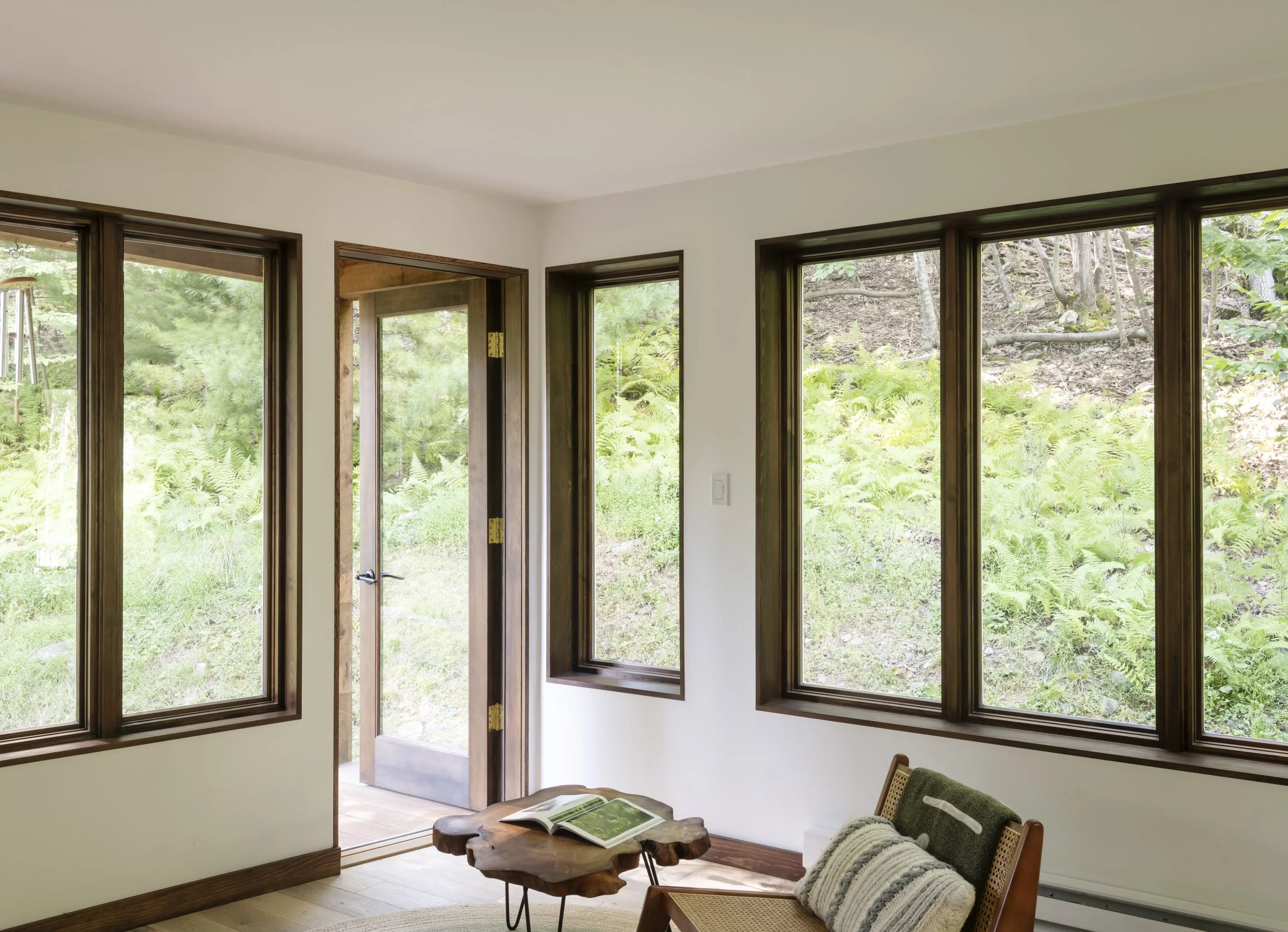 Interior view of a room with large windows and a view of a lush, green outdoor area with trees and plants, a wooden door, a small wooden coffee table with an open book, and a chair with a blanket.