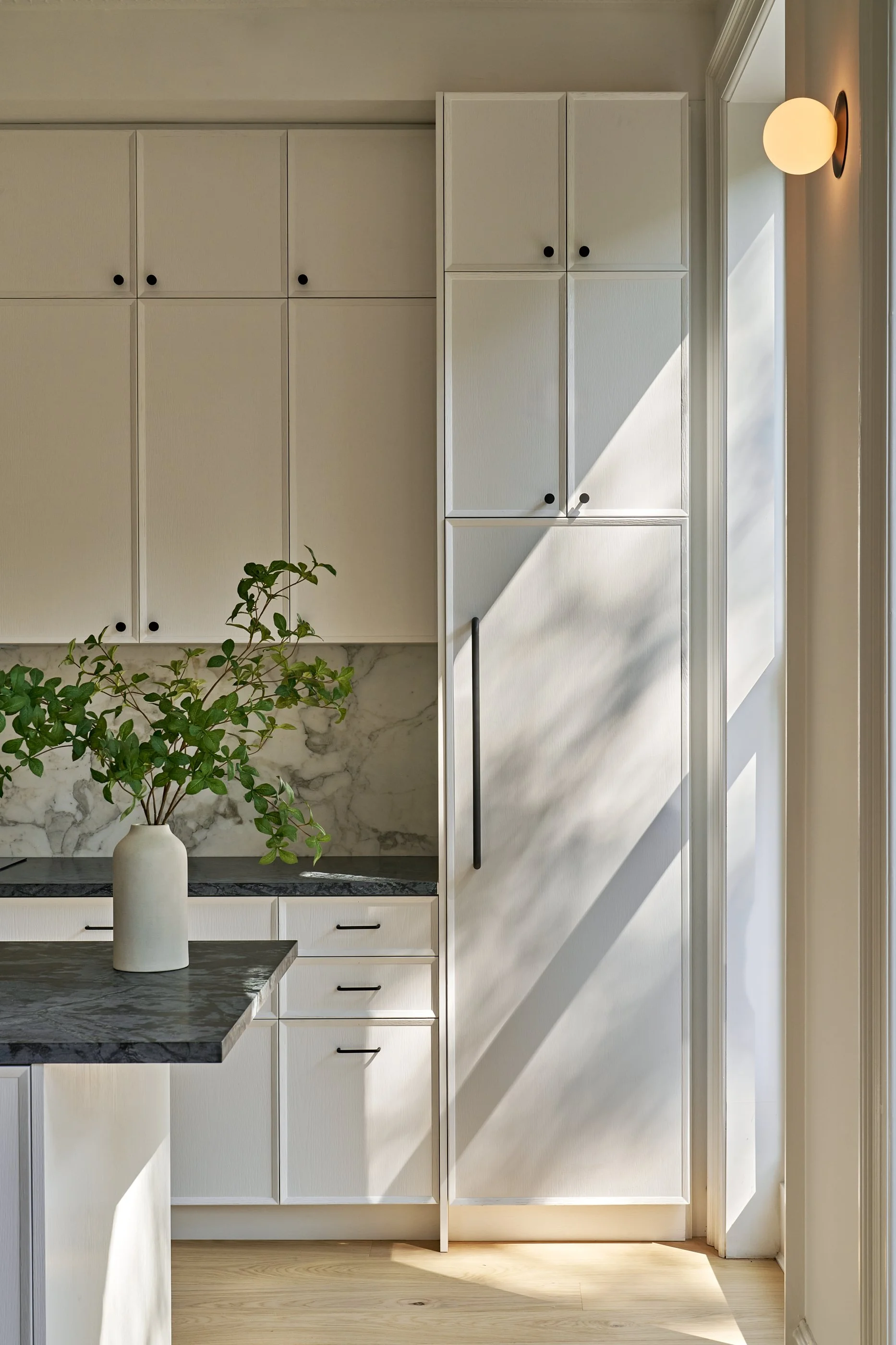 Interior view of a modern kitchen with white cabinets, a marble backsplash, and a black handle on a tall cabinet, illuminated by natural sunlight and a wall-mounted light fixture.
