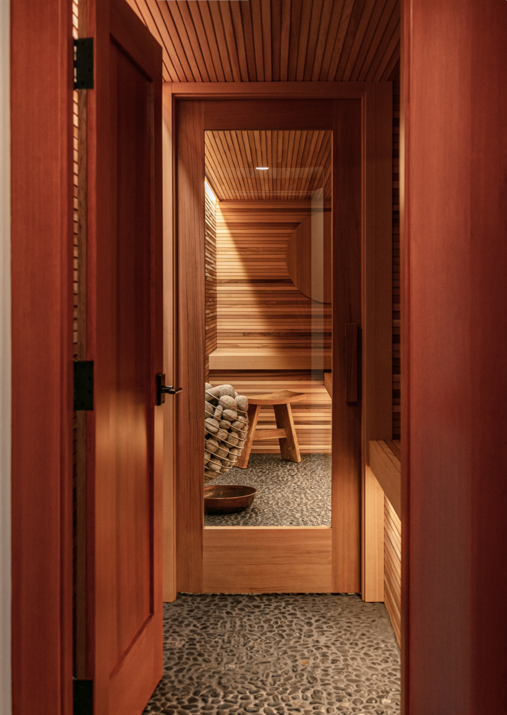 Their sauna room view with wooden panels, a basket filled with sauna stones, a small wooden bench, and a textured pebble floor. Victoria British Columbia Custom Home. Architecture and Interior Design.