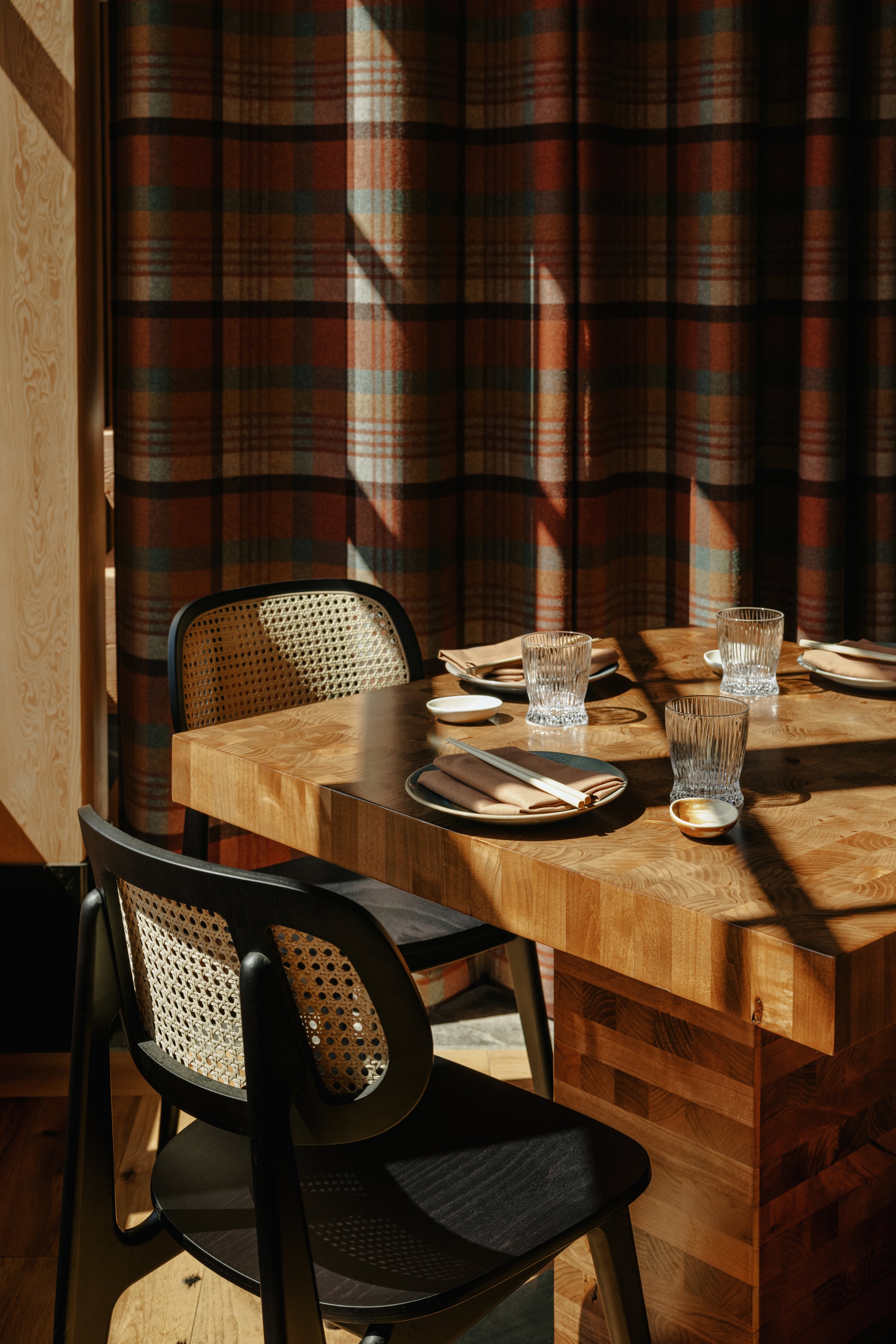 A wooden dining table set with glasses, plates, and napkins, with sunlight casting shadows on a plaid curtain in the background. Hello Sunshine Banff Alberta. Architecture and Interior Design.
