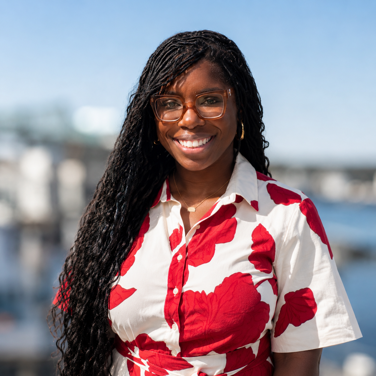 Smiling woman with long black braids and glasses, wearing a white dress with red floral pattern, standing outdoors by water with bridge in background.