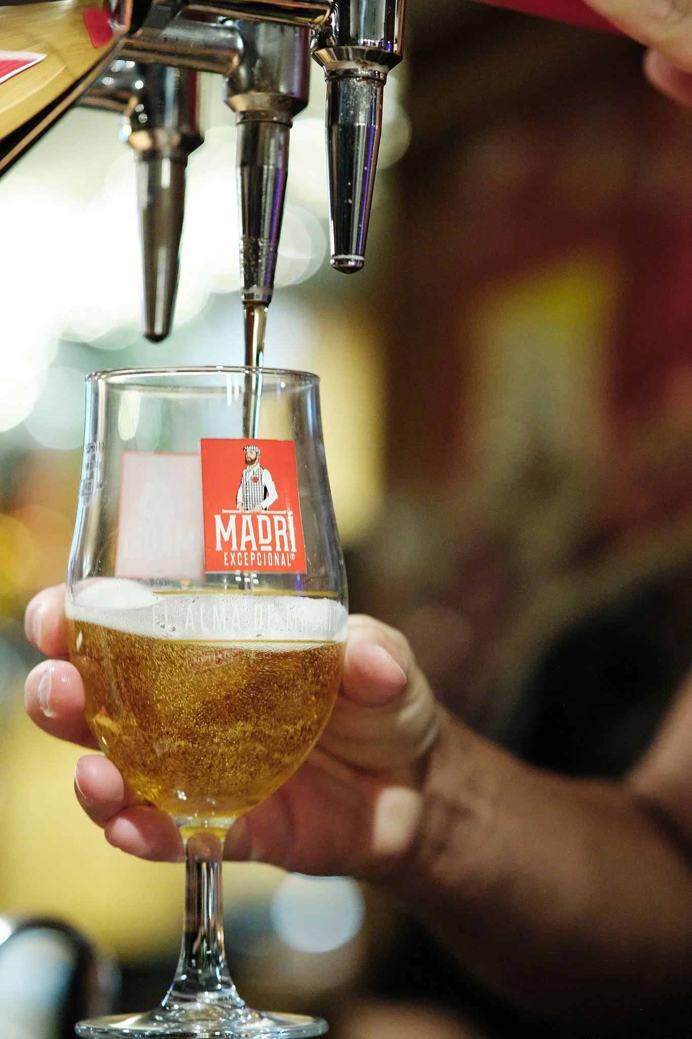 A hand holding a glass of beer under a beer tap at a bar. The glass has a red sticker with a chef and the words 'MADRI EXCEPCIONAL' on it.