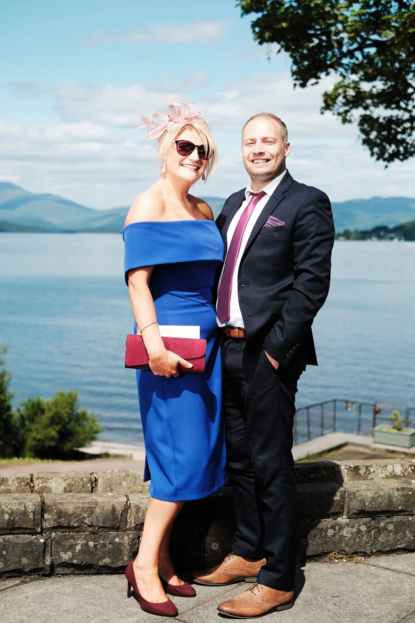 A smiling woman in a blue off-shoulder dress and maroon heels standing next to a smiling man in a dark suit and tie, near a body of water, with mountains and a tree in the background.