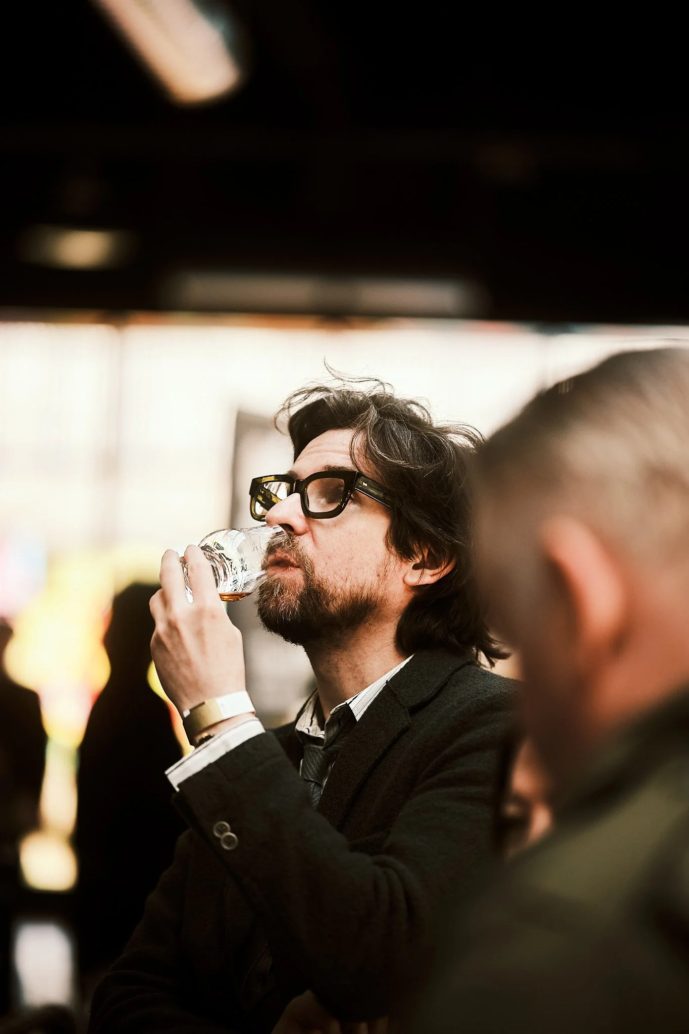 A man with glasses and beard drinking from a small glass in a crowded indoor setting.