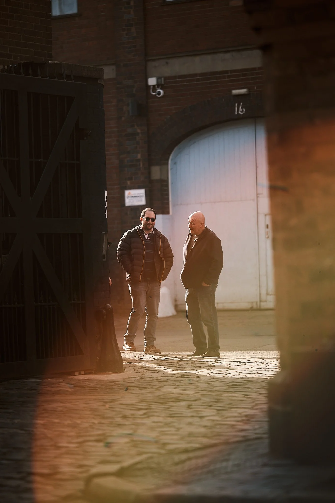 Two men standing and talking in an outdoor urban area near a large white gate, with one wearing sunglasses and a jacket, and the other in a black blazer and jeans.