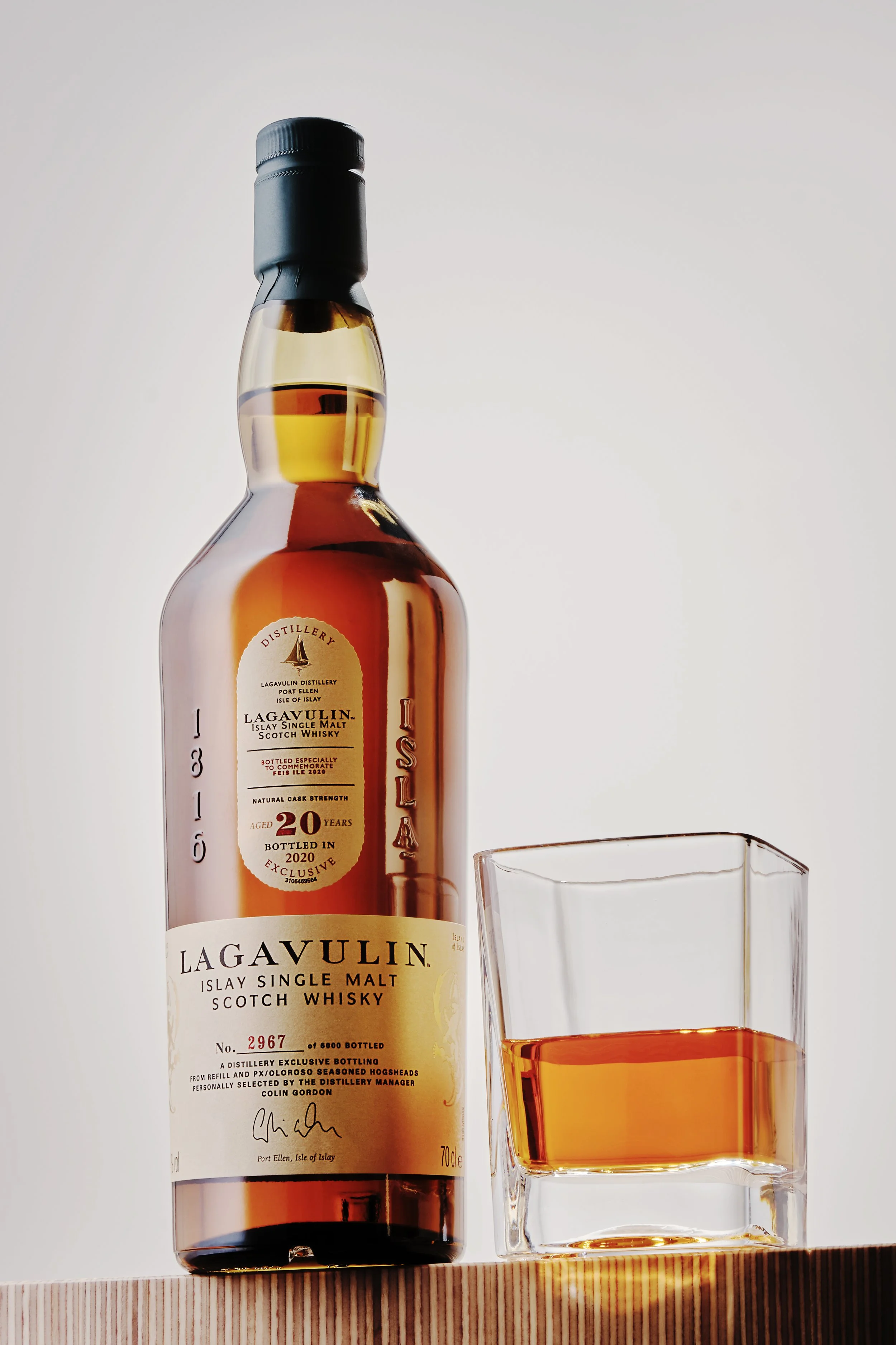 A bottle of Lagavulin 20-year-old single malt Scotch whisky next to a glass filled with whisky on a wooden surface against a plain background.