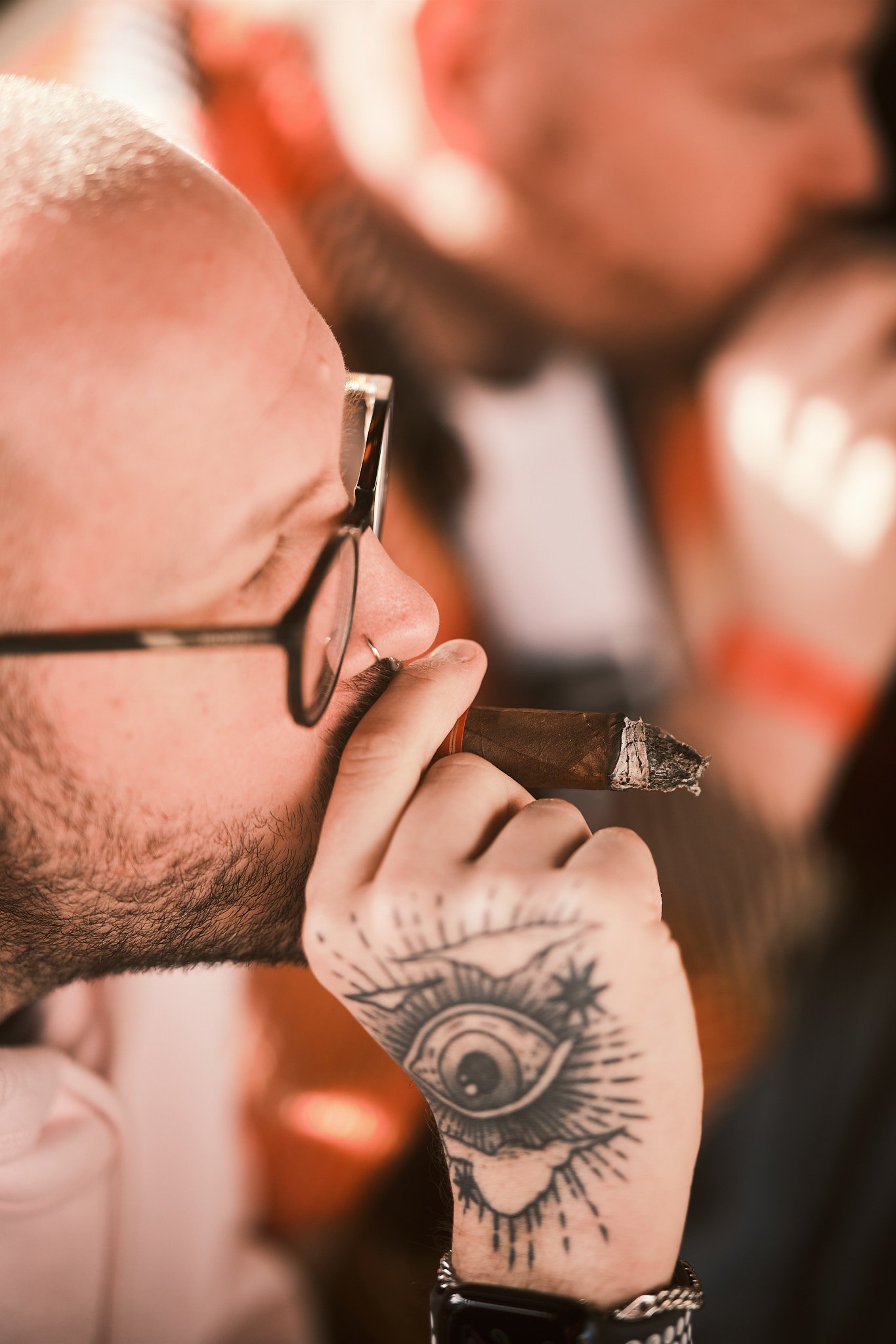 Close-up of a man with glasses and a nose piercing smoking a cigar, showing a tattooed hand with an eye and star design.