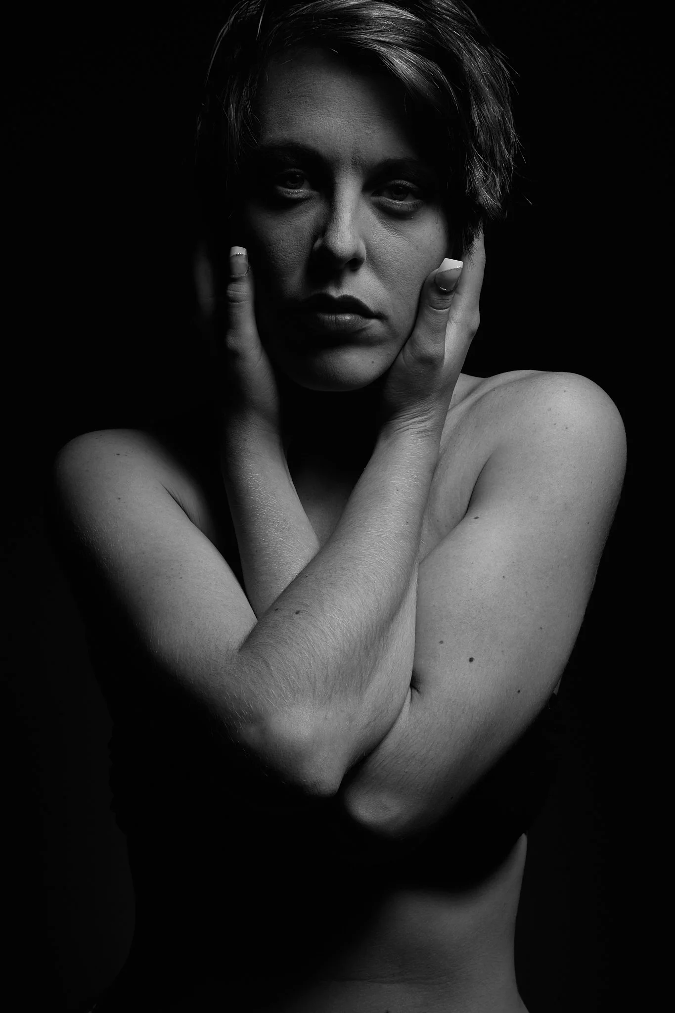 Black and white portrait of a woman touching her face and shoulders with her hands, looking directly at the camera against a dark background.