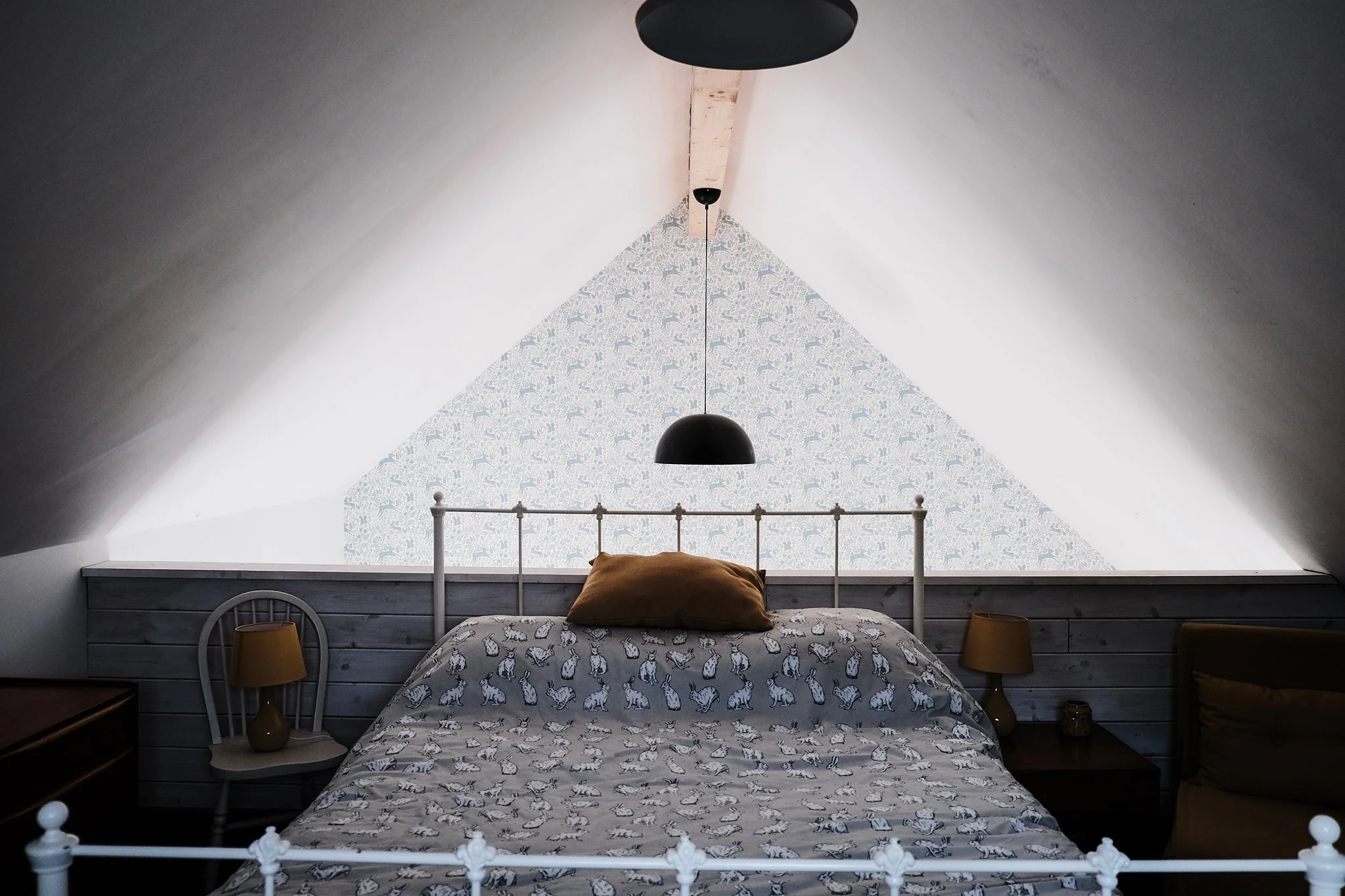 Bedroom with white metal bed frame, patterned bedding with rabbits, brown pillow, wooden nightstands with lamps, slanted ceiling with patterned wallpaper behind the bed, black pendant light hanging from ceiling