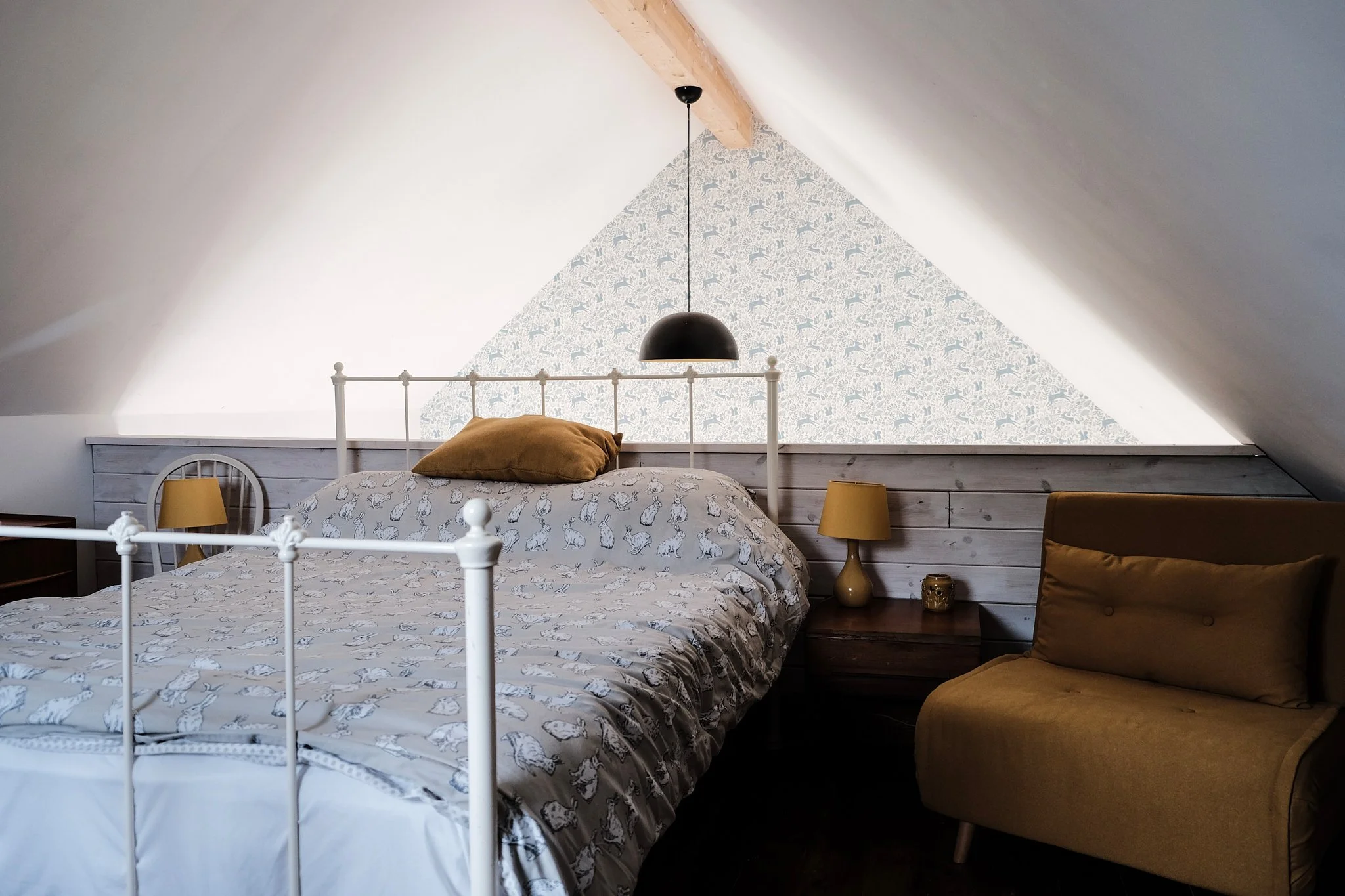 Bedroom with sloped ceiling, white metal bed frame, patterned bedding, brown pillows, yellow lamps on nightstands, wooden beams, and a mustard yellow sofa.