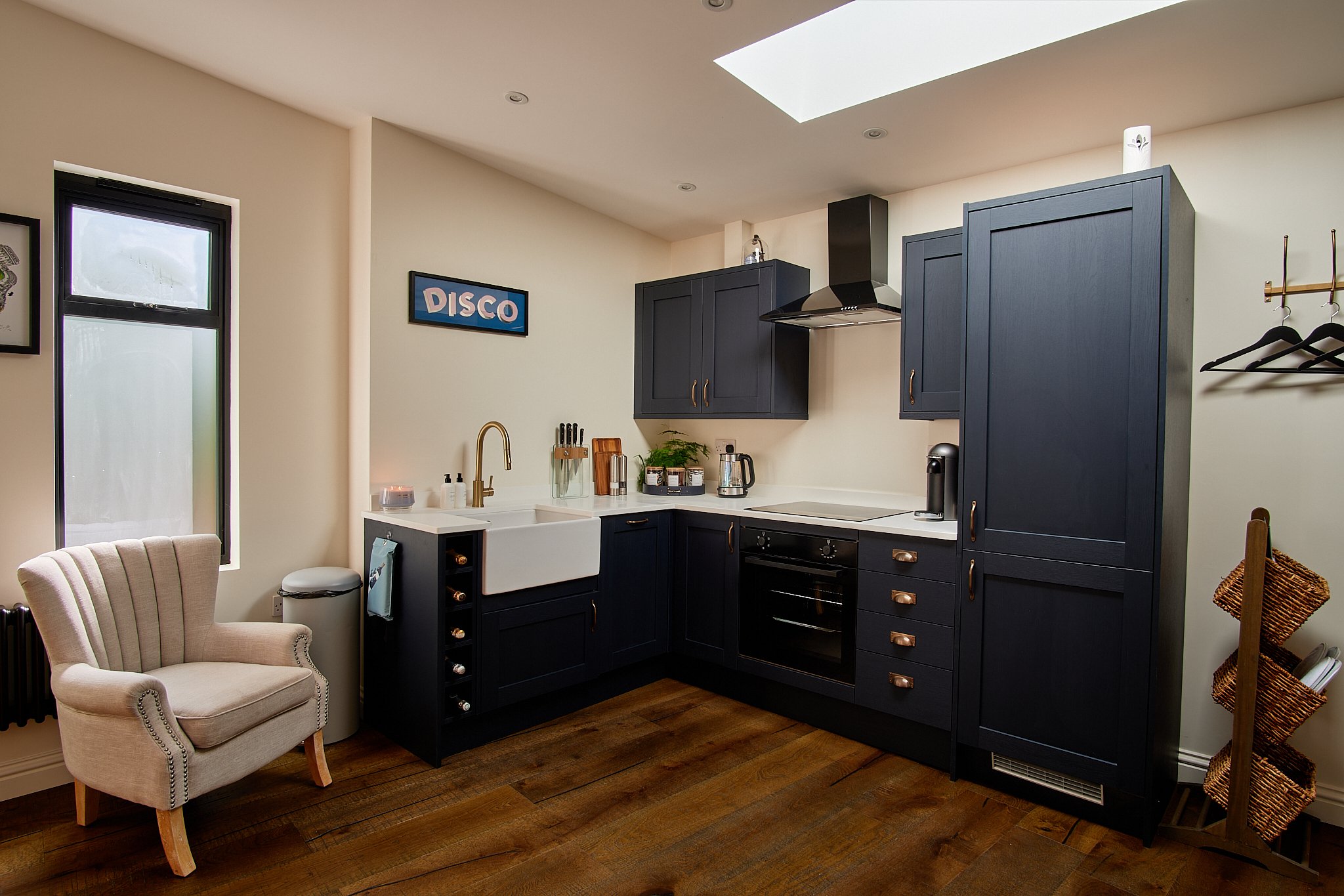 Modern kitchen with blue cabinetry, white countertops, and a farmhouse sink. There is a chair in the corner, a window, and a wall sign that says 'DISCO'. A wooden wine rack and woven baskets are also visible.