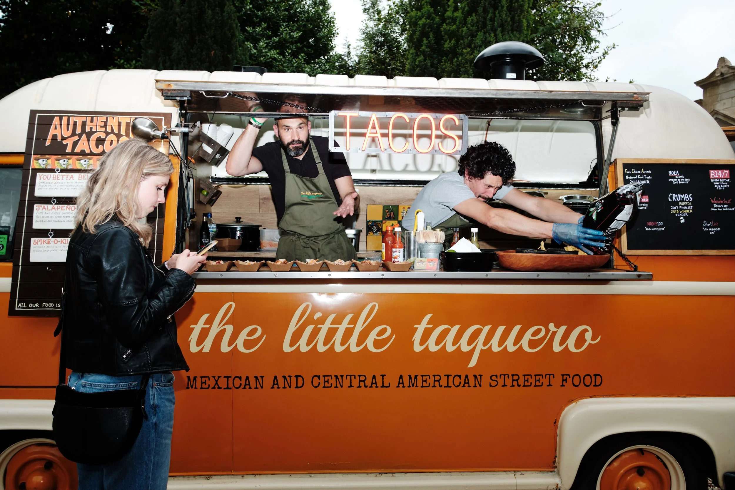 A food truck named 'the little taquero' serving Mexican and Central American street food, with two male staff preparing food and a young woman in a black leather jacket looking at her phone.