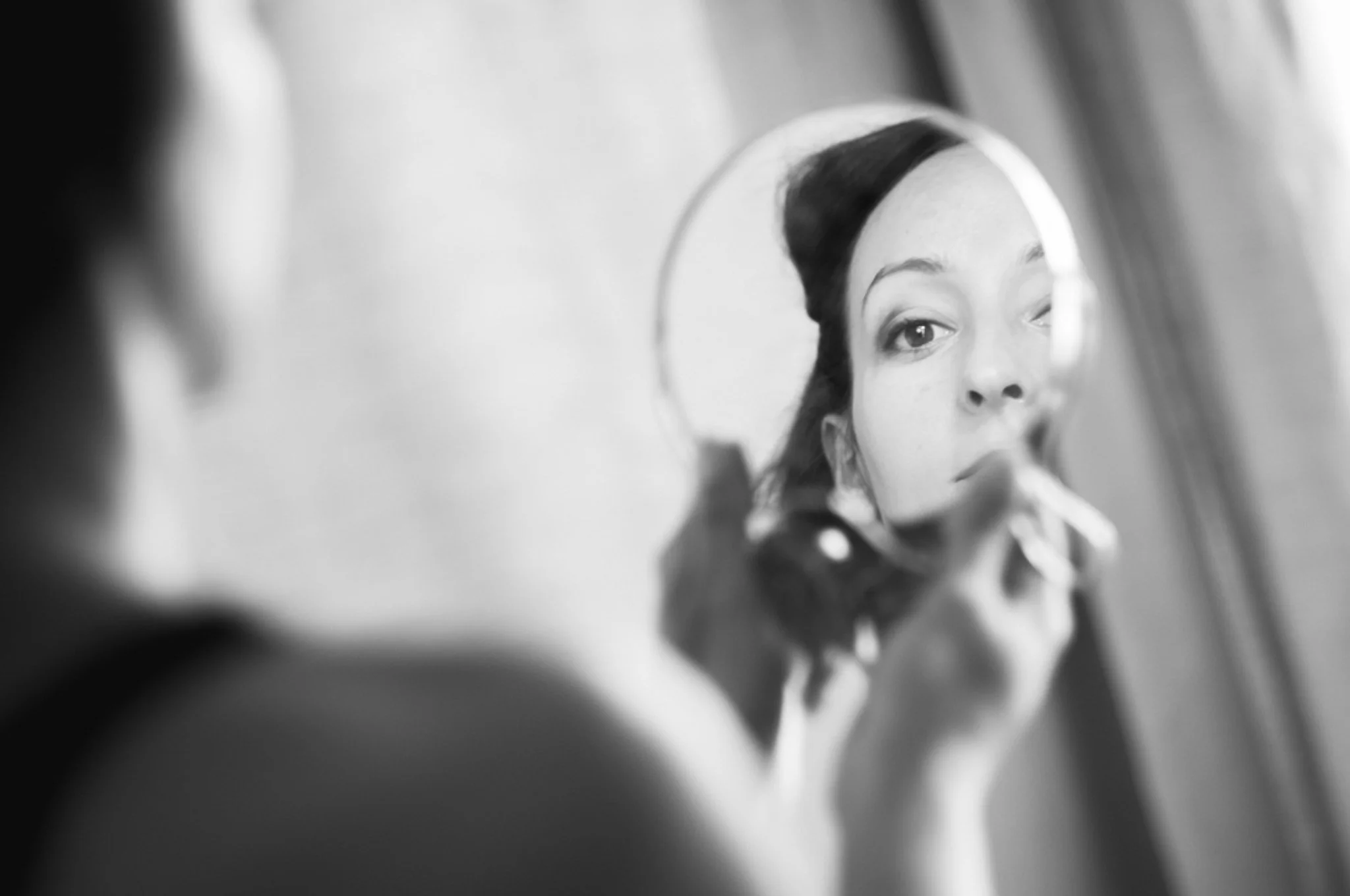 A person with braids looking into a mirror, their face reflected in the mirror with a neutral expression. The image is in black and white.