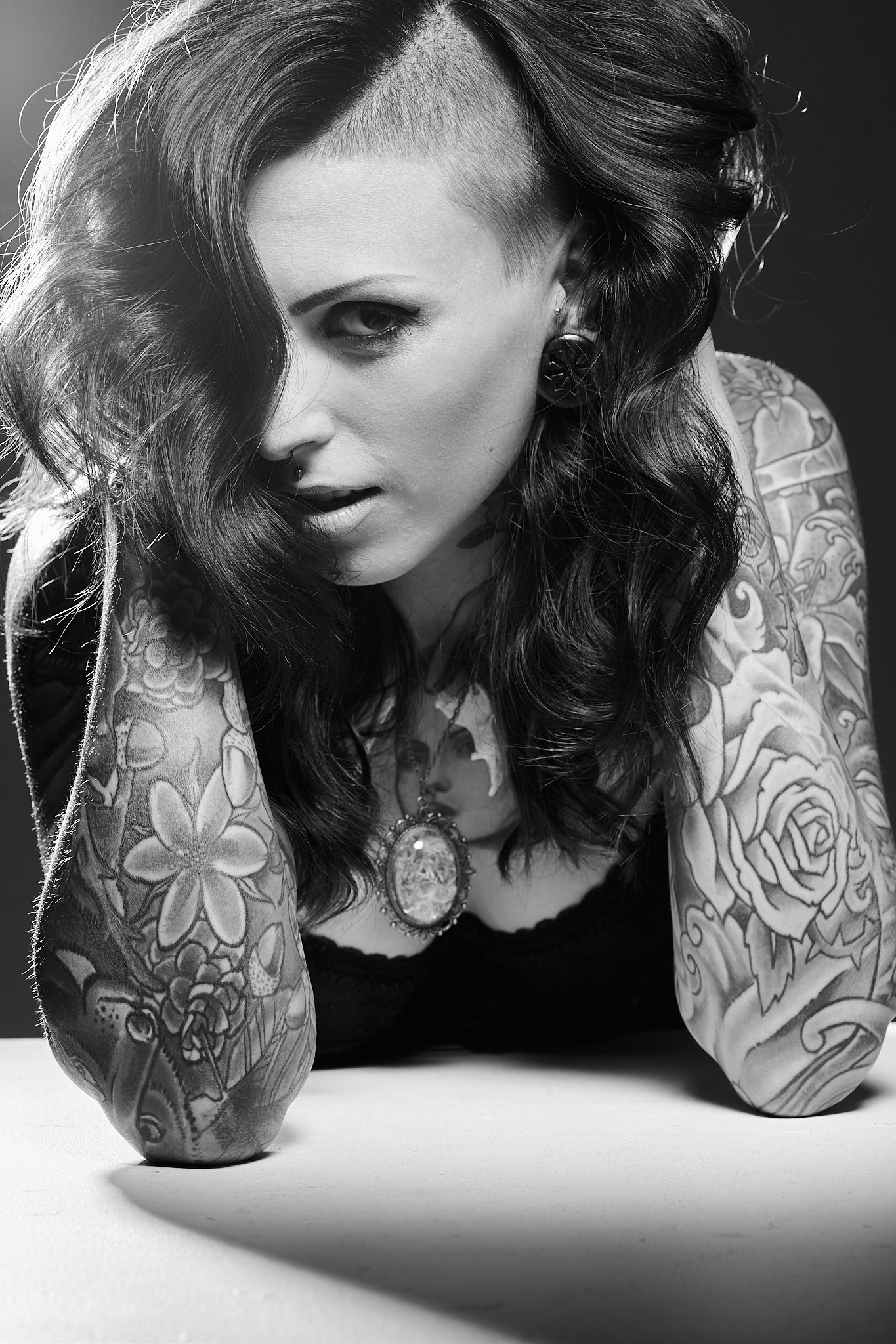 A woman with tattoos on her arms and chest, dark wavy hair, and piercings, looking intensely at the camera with one eye visible, in black and white.