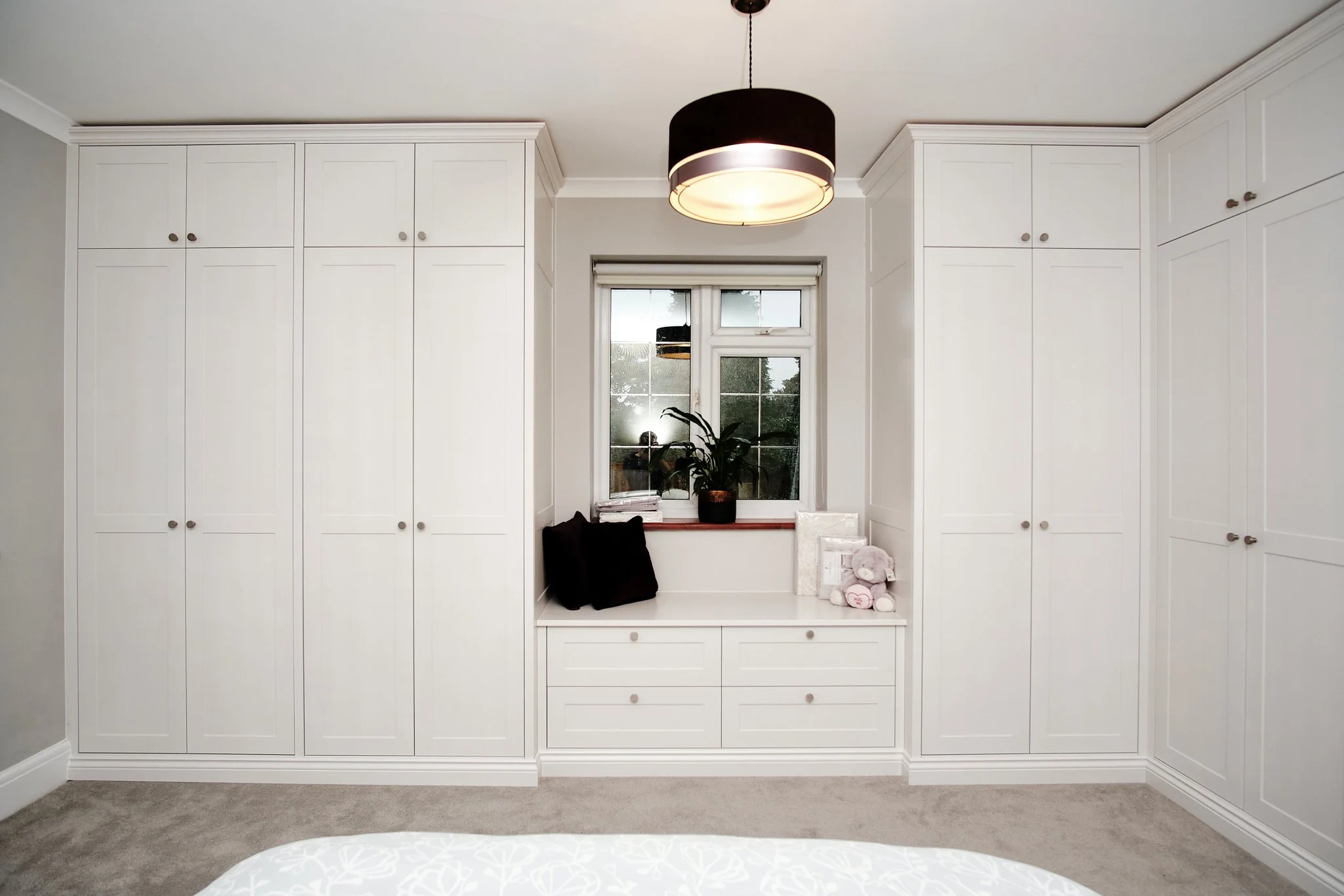 White built-in cabinets and drawers surrounding a window with a black light fixture hanging from the ceiling, a potted plant, and some decorative pillows and stuffed animals on a built-in bench below the window.
