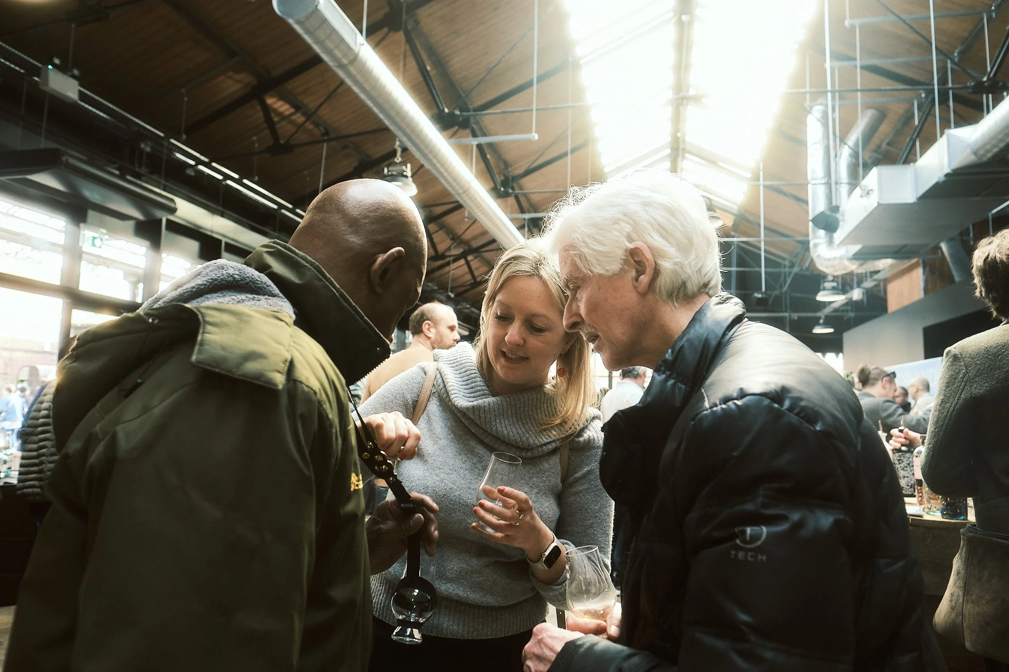 Three people engaged in conversation at an indoor event, two men and one woman, with glasses of wine in their hands. The setting appears to be a modern, industrial-style venue with natural light coming through skylights.