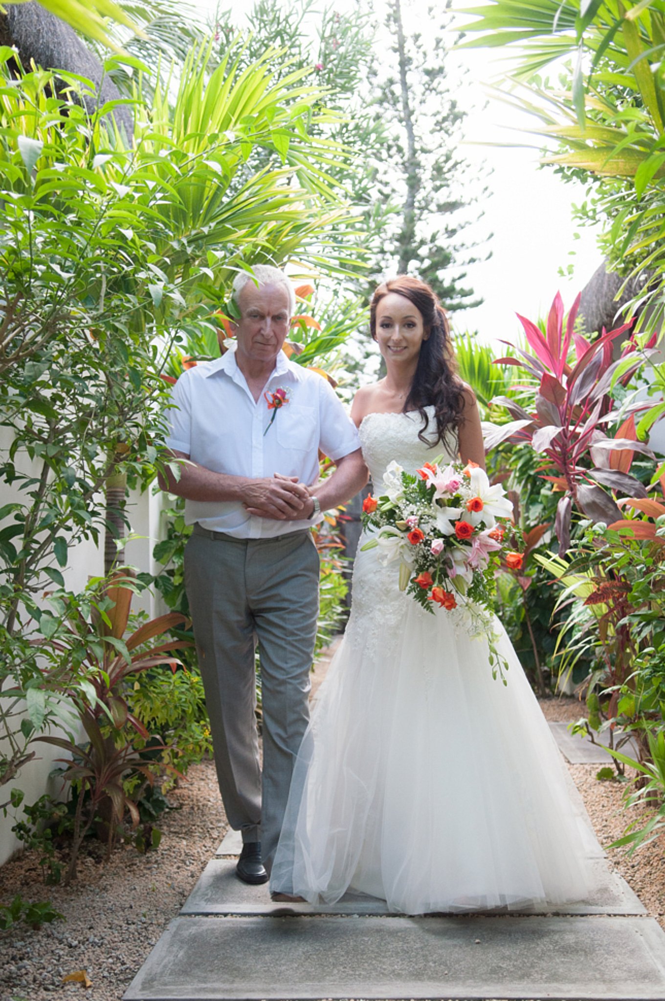 A bride and an older man, possibly her father, standing together in a lush, green garden with tropical plants. The bride is holding a colorful bouquet and wearing a white wedding dress. The man is dressed in a white shirt and gray trousers, and they 