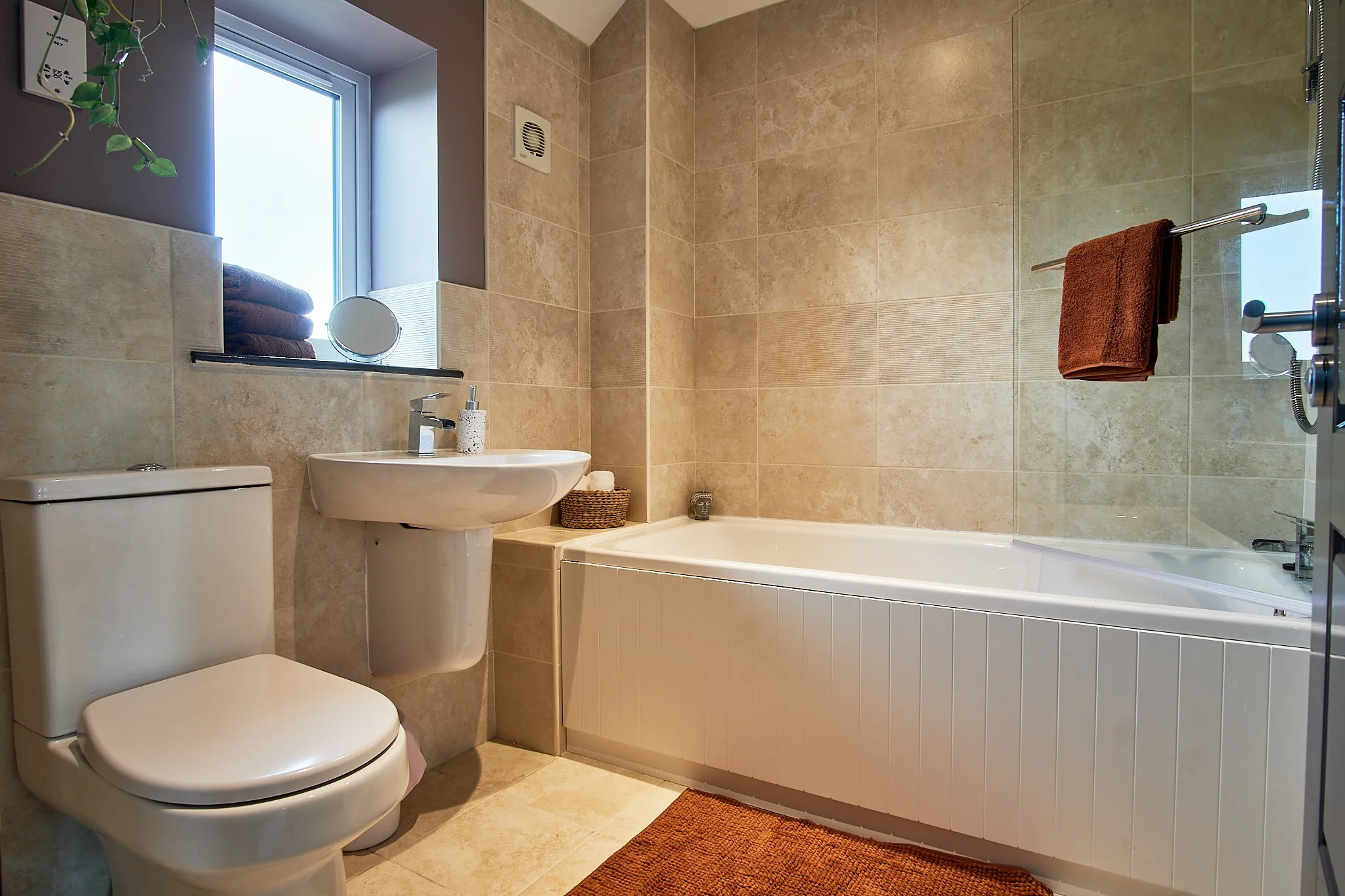 Bathroom with beige tiled walls and floor, a white toilet, a small wall-mounted white sink with a silver faucet, a small basket next to the bathtub, a window with a ledge holding folded towels and a round mirror, and a brown towel hanging on a towel 