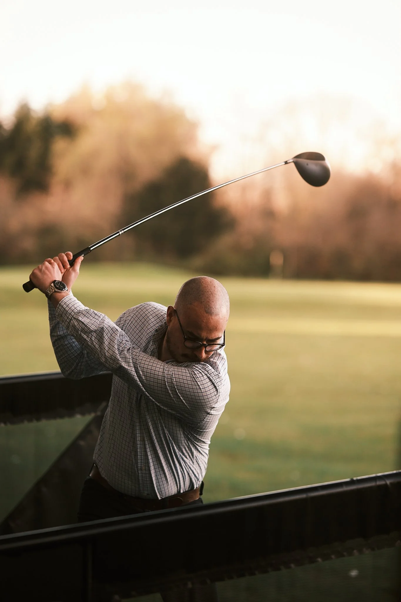 Man hitting golf ball on golf course, mid-swing, wearing glasses and checkered shirt, with a blurred background of trees and grass.