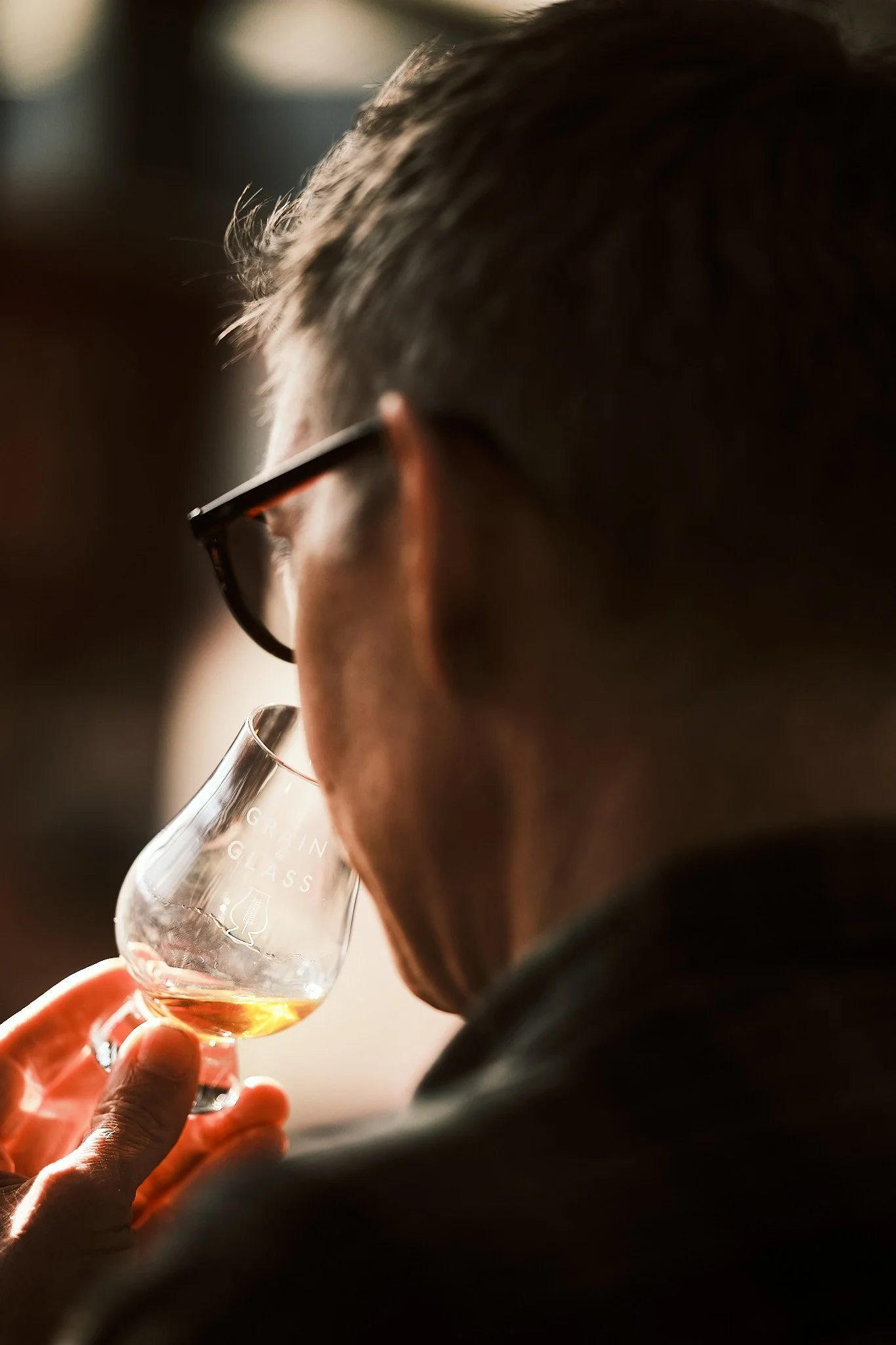 Person drinking a glass of whisky or bourbon, with 'GRAIN GLASS' inscribed on the glass, in a close-up side profile view.