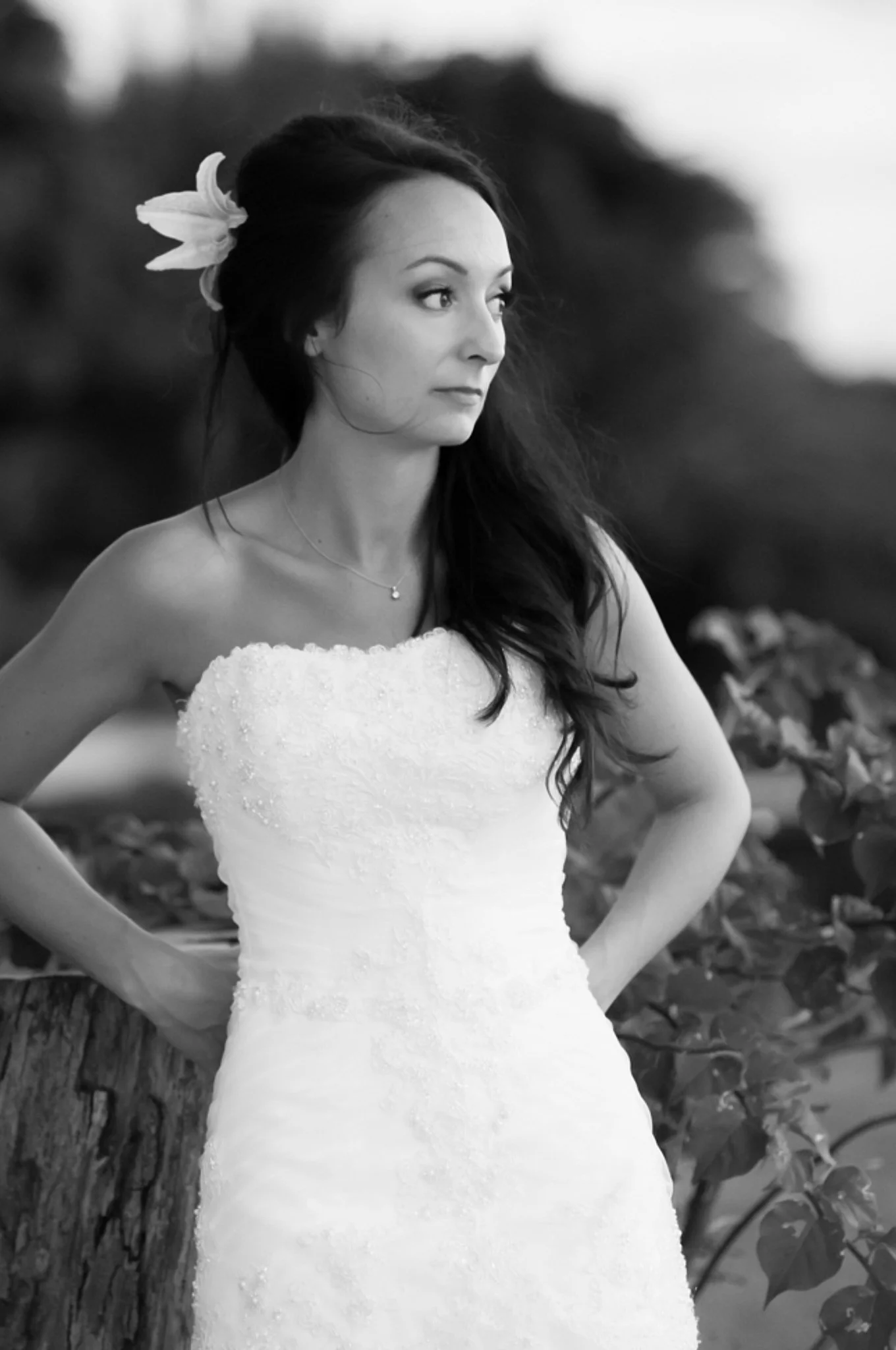 Black and white photo of a woman in a strapless dress with a flower in her hair standing outdoors by a tree.