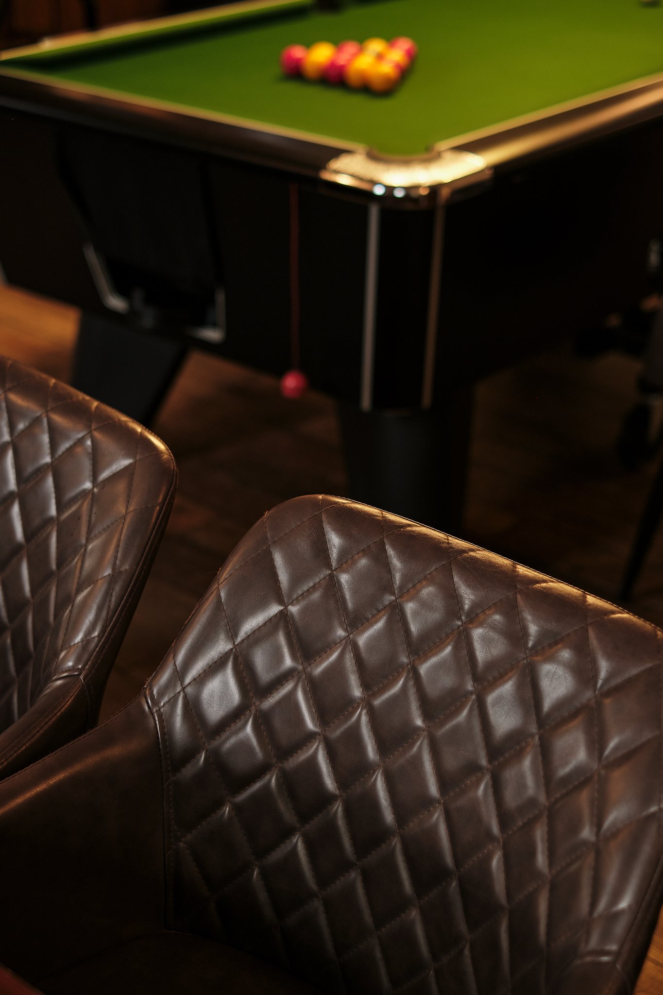 Close-up of brown leather chairs with quilted pattern in front of a pool table with billiard balls arranged for a game.