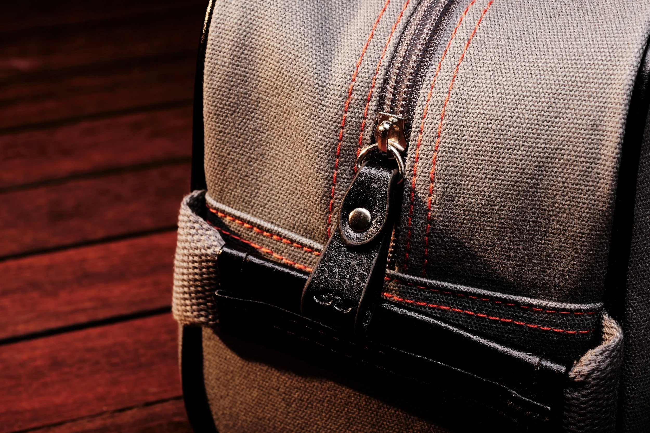 Close-up of a gray backpack with a zipper pocket and black leather zipper pull, set against a wooden background.