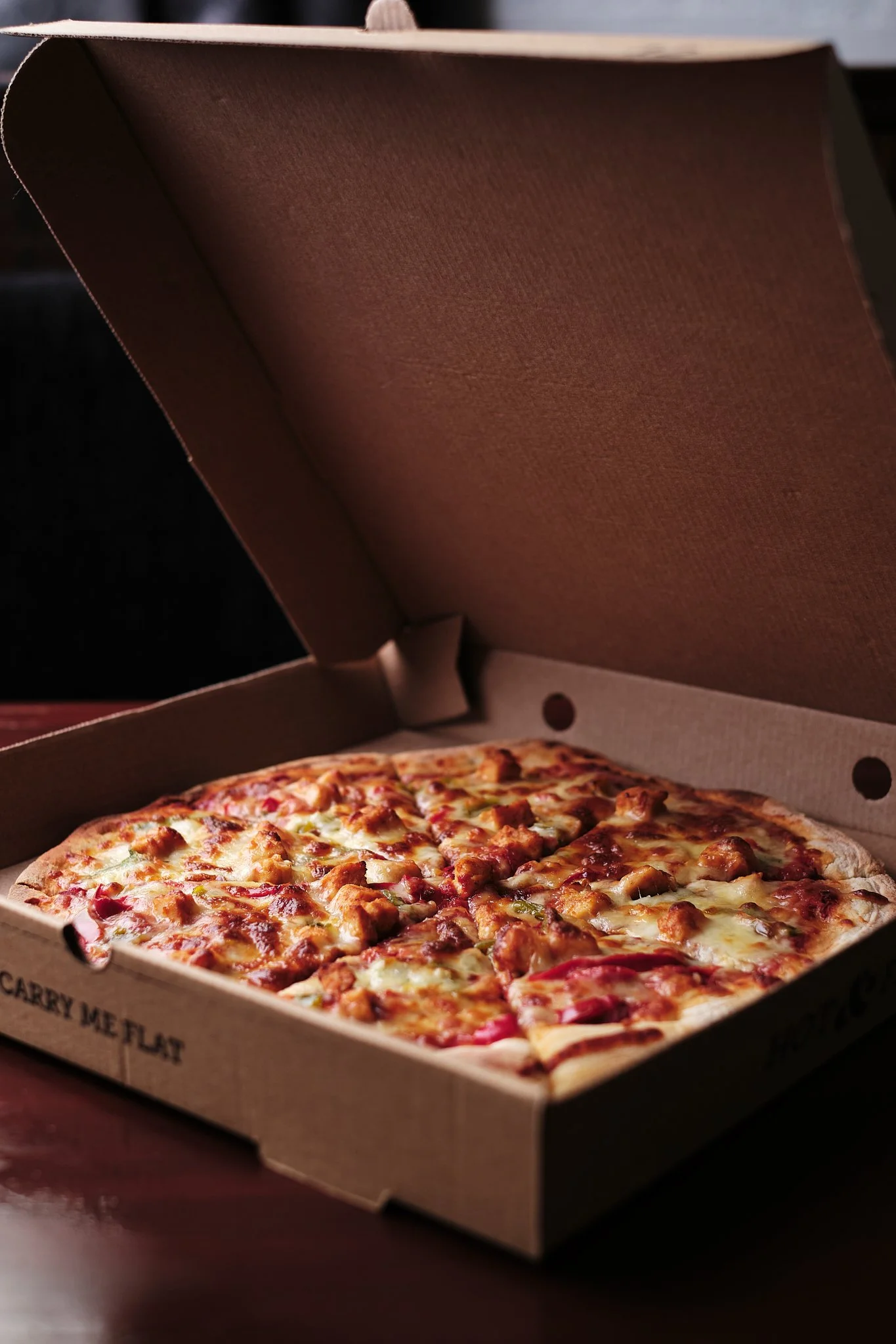 A pizza in an open cardboard takeout box with chicken, cheese, and toppings inside.