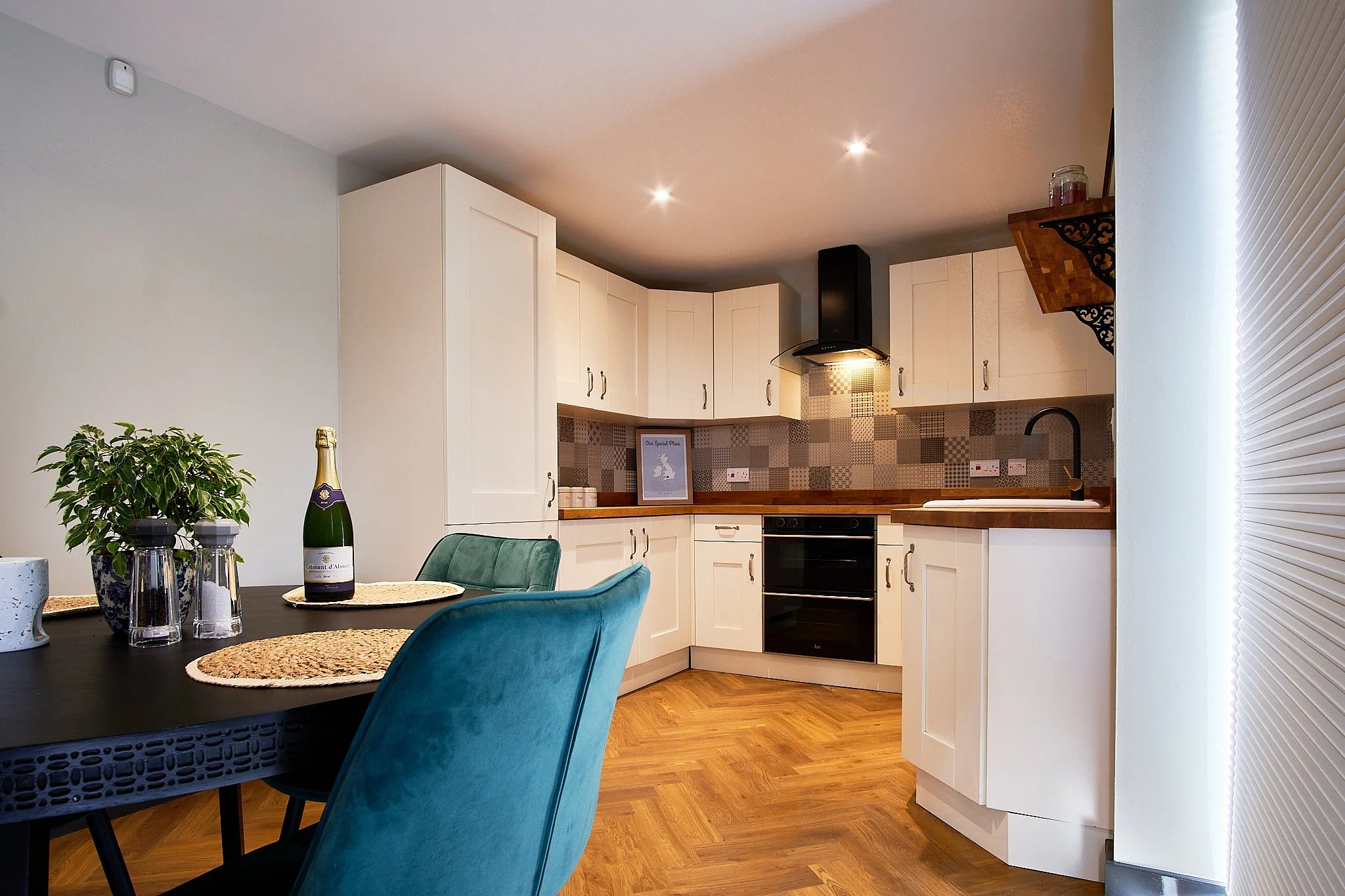 Modern kitchen with white cabinets, black oven, wooden countertops, and a tiled backsplash. Dining table with colorful chairs and decorative placemats, and a potted plant on the table.