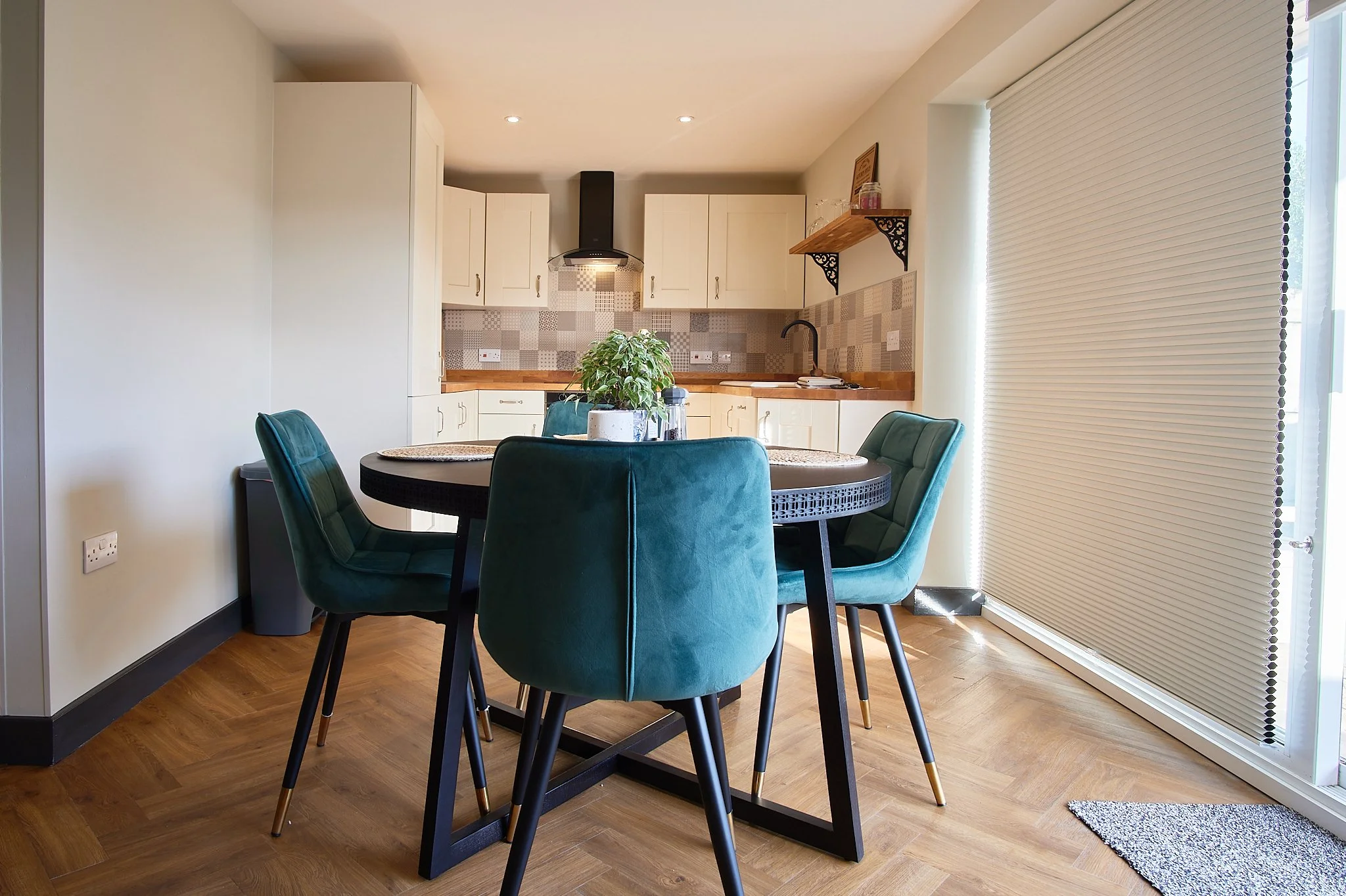 Modern dining area with a round black table, four green velvet chairs, a potted plant in the center, wooden flooring, and a kitchen with white cabinets, a patterned tile backsplash, and a large window with closed blinds.