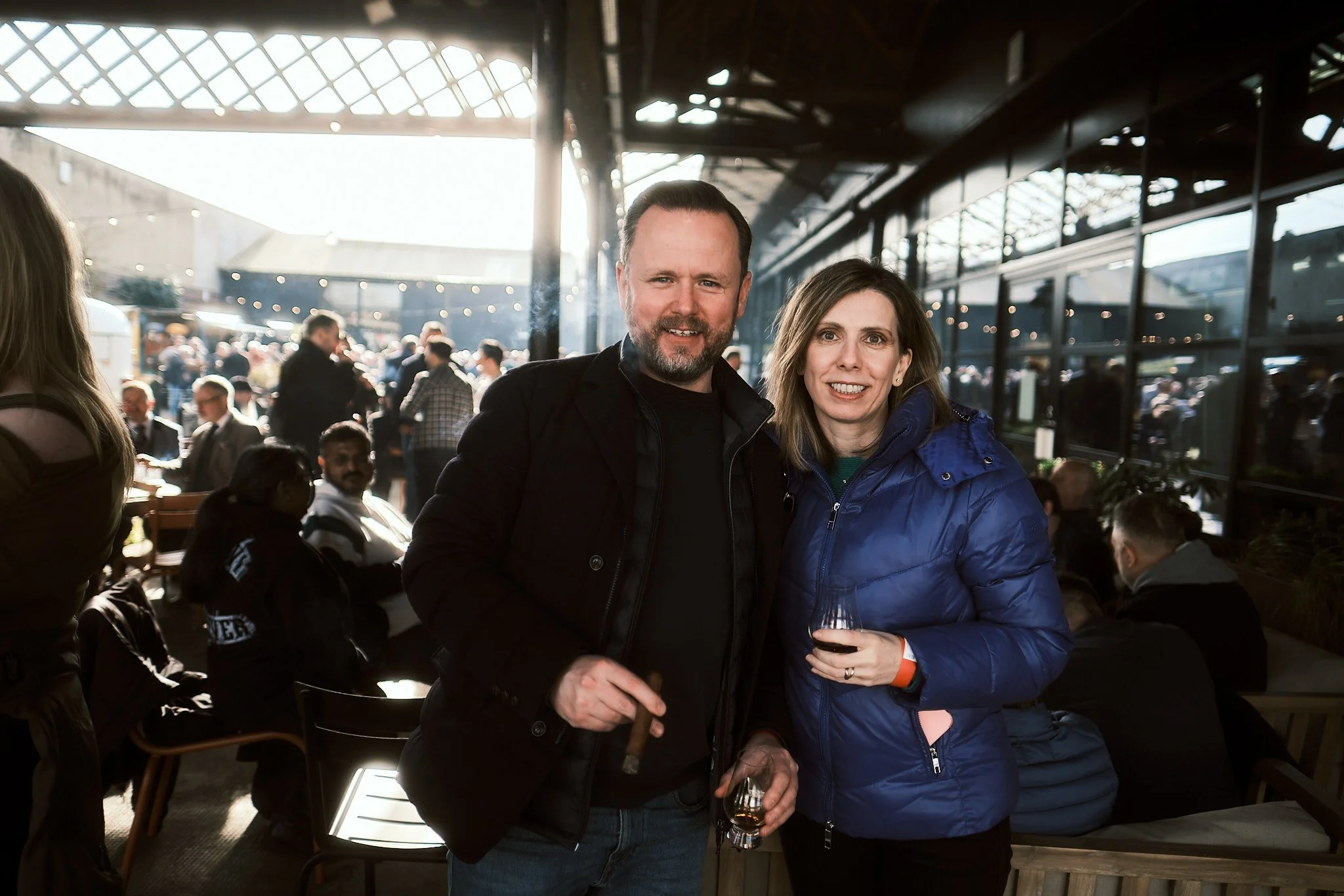 A man and a woman standing together at a social gathering, both holding drinks, with a crowd of people in the background at a well-lit indoor-outdoor venue.