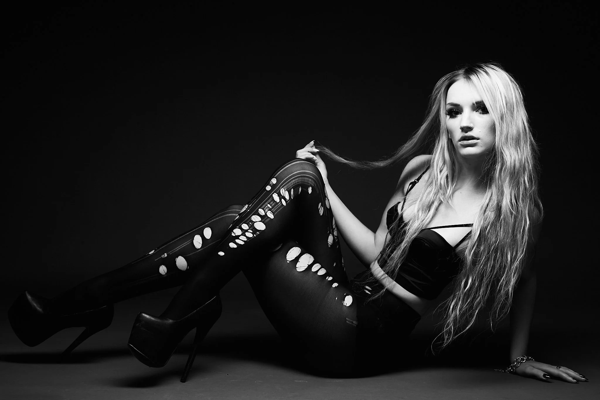 Black and white photo of a young woman with long wavy hair, wearing high-heeled boots with circular cutouts, a black top, and ripped tights, sitting on the floor against a dark background.