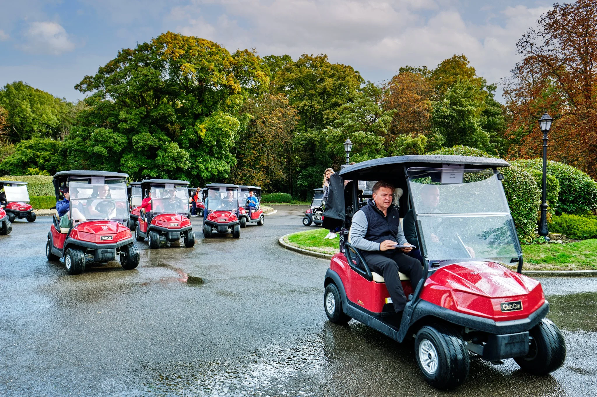 Group of people riding red golf carts on a wet driveway, surrounded by green trees and bushes, with street lamps and cloudy sky in the background.