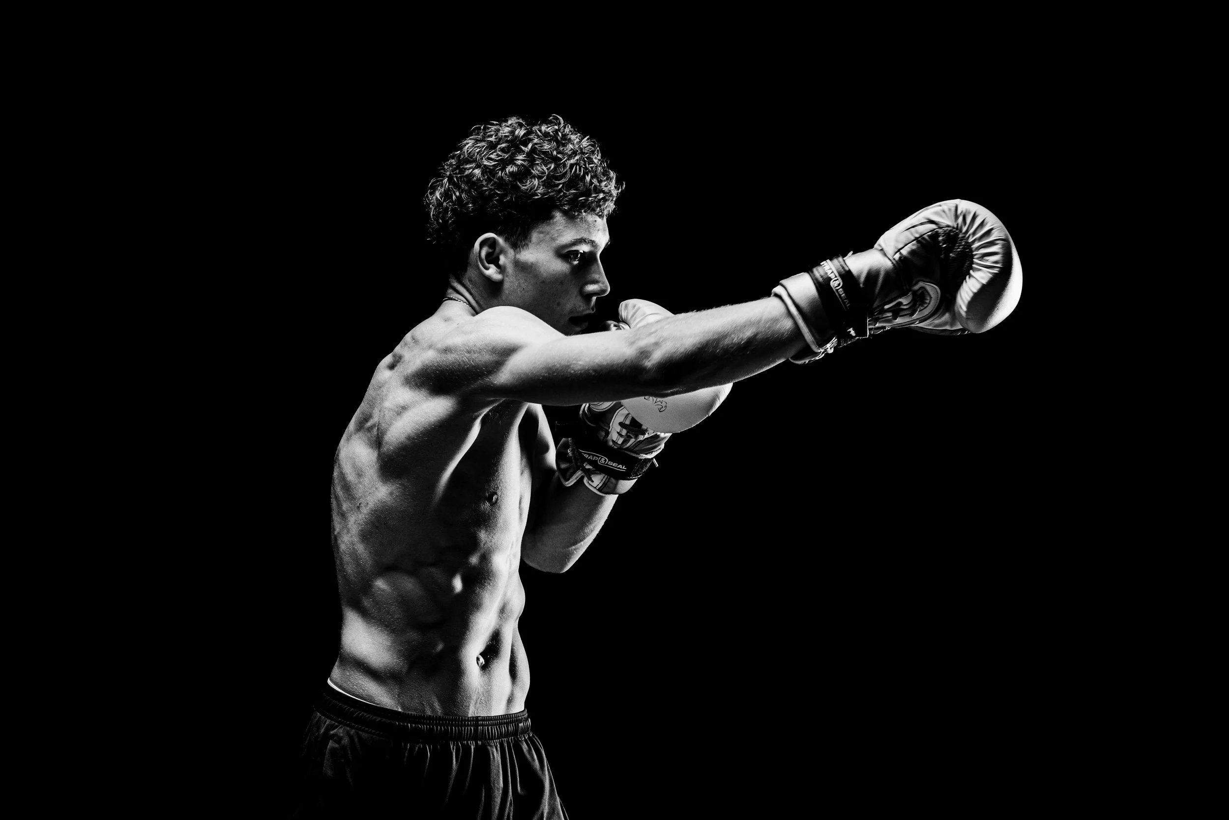 Black and white photo of a young male boxer with curly hair, shirtless, wearing boxing gloves, in a boxing stance against a black background.