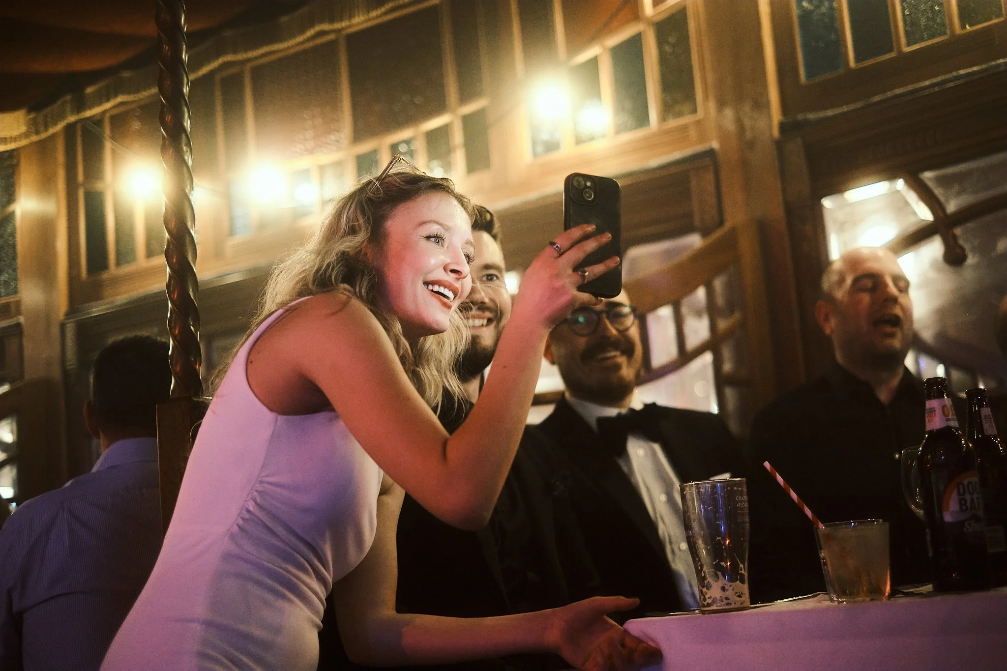 A young woman in a white dress taking a selfie at a party or celebration, surrounded by smiling men dressed in formal attire, inside a warmly lit wooden venue with decorative glass windows.