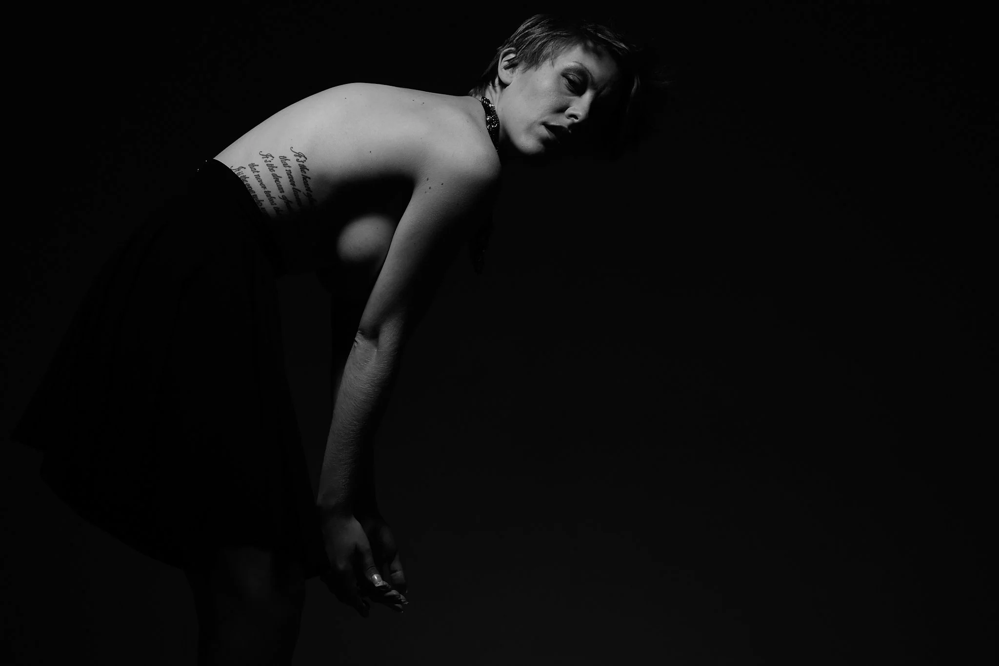 A black and white photo of a woman with short hair, wearing a necklace, bent forward with her left arm hanging down and her right arm bent and resting on her knee, against a dark background.