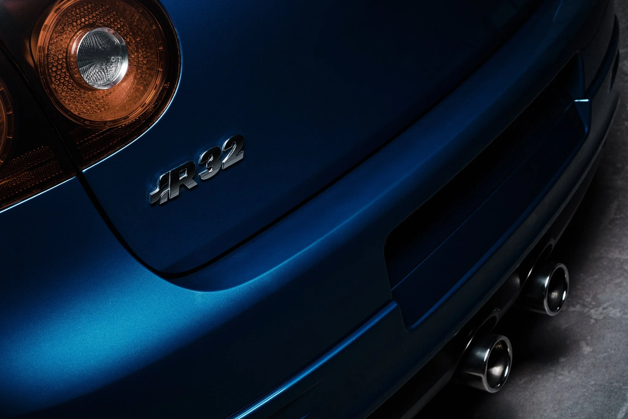 Close-up of the rear of a blue sports car, showing the taillight, model badge 'R32', and dual exhaust pipes.