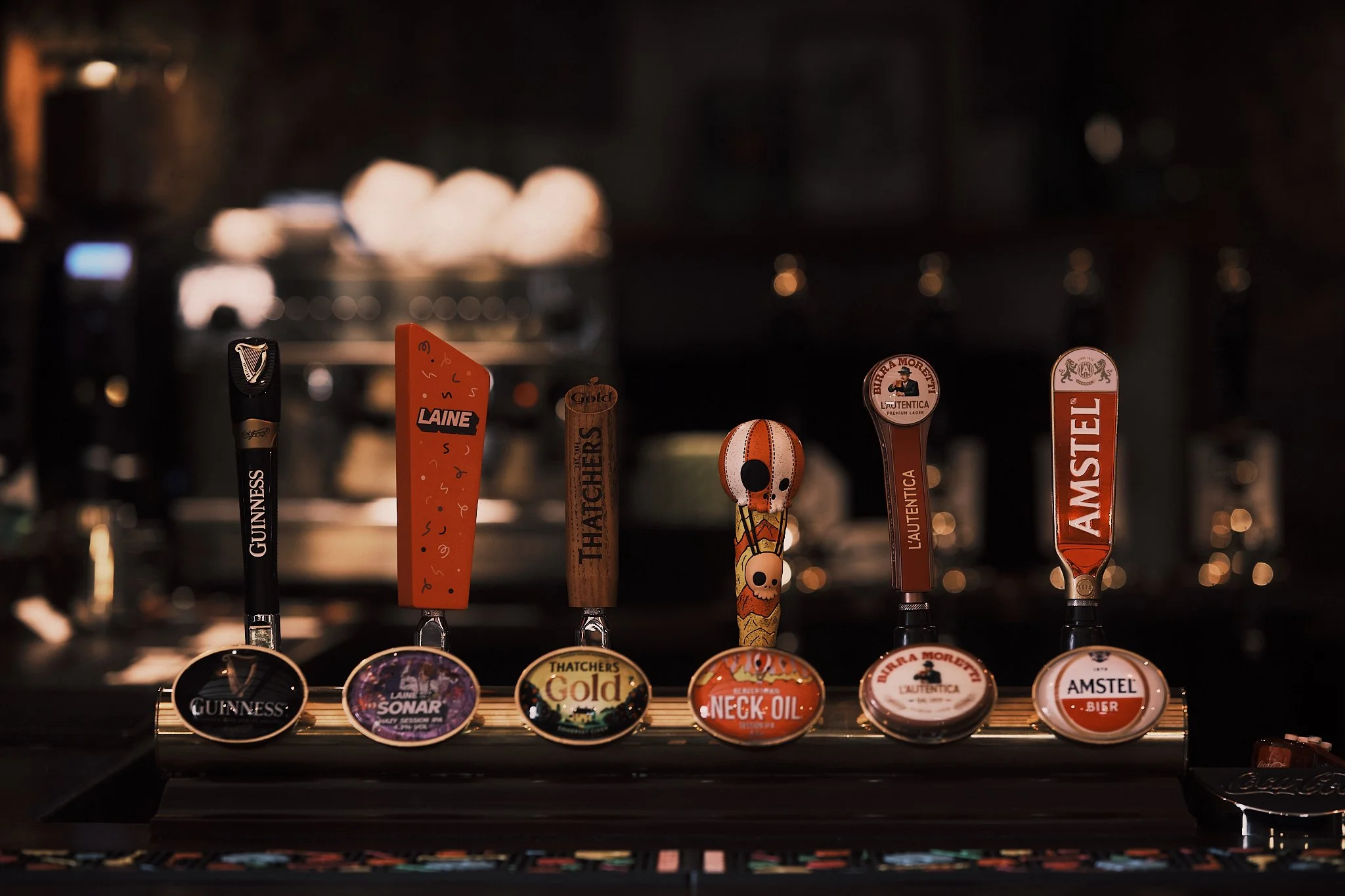Six beer tap handles at a bar, each with a different brand, including Guinness, Laine, Thatchers, Neck Oil, La Tента, and Amstel, with the labels visible on the tap handles.