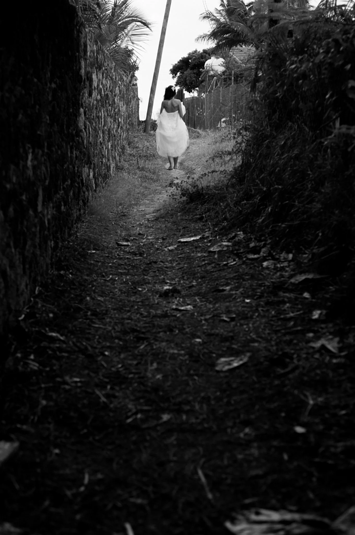 A woman wearing a white dress walking down a narrow dirt path between stone walls and surrounded by trees and bushes.